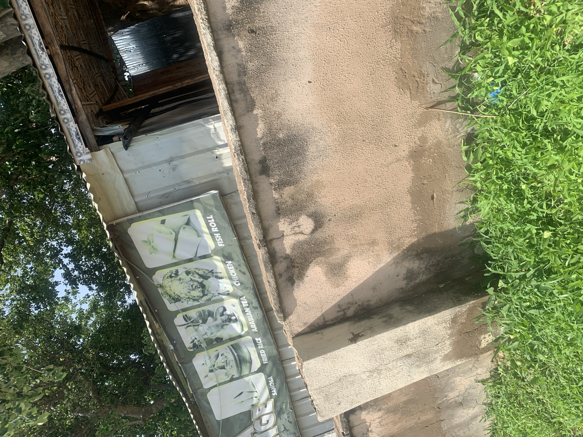 The image shows an outdoor scene in Jalingo, Nigeria. A corrugated metal building with a decorative trim along the top edge is visible. A banner displaying images of food items such as samosas, fried rice, Arabian tea, chicken, and fish rolls is affixed to the front of the building. A rough concrete wall, stained and weathered, runs along the lower portion of the image, with lush green grass growing at its base. The scene suggests a food stall or small eatery in a somewhat unkempt urban environment.