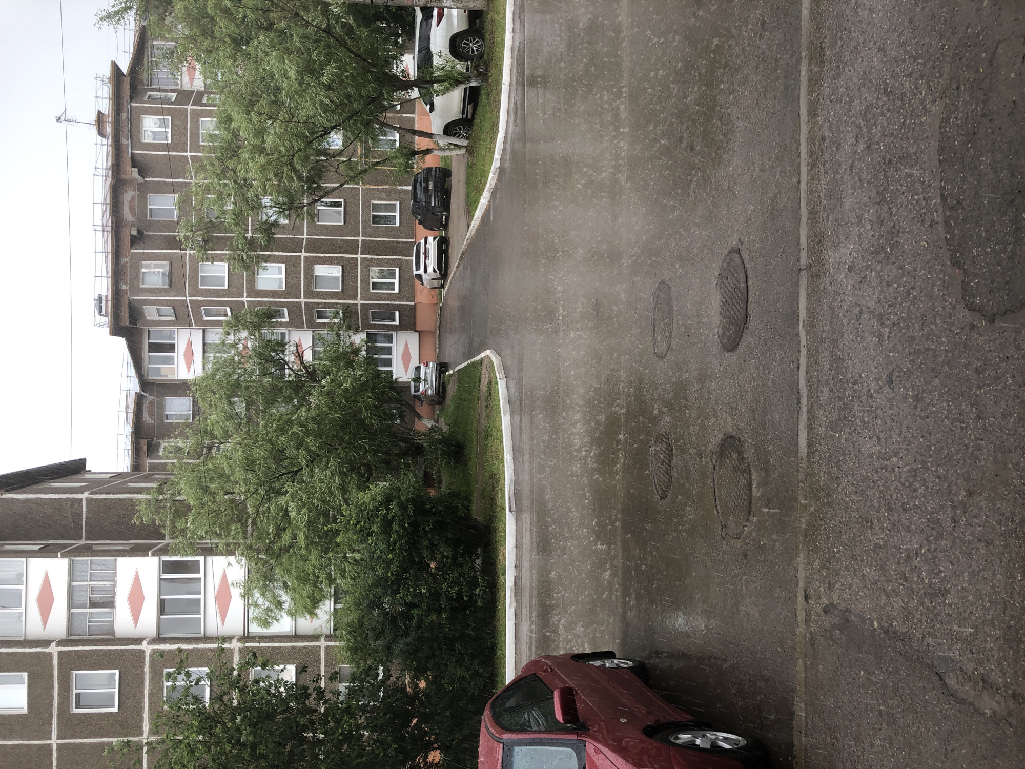 This media file depicts an outdoor scene on a cloudy day, likely in Solikamsk, Russia, as suggested by the context. The setting is a residential area featuring a multi-story concrete building with numerous square windows. In front of the building, several cars are parked along a wet asphalt road, indicating it is raining or has recently rained. Trees with green foliage are visible, some of which are close to the building, obscuring parts of the facade. The road surface is reflective, showing the effects of the rain, and there are at least two manhole covers visible on the pavement. A red car is parked in the foreground, partially obscuring a grassy area and a sidewalk. The overall mood is subdued, consistent with overcast and rainy weather. There is no visible text in the image.