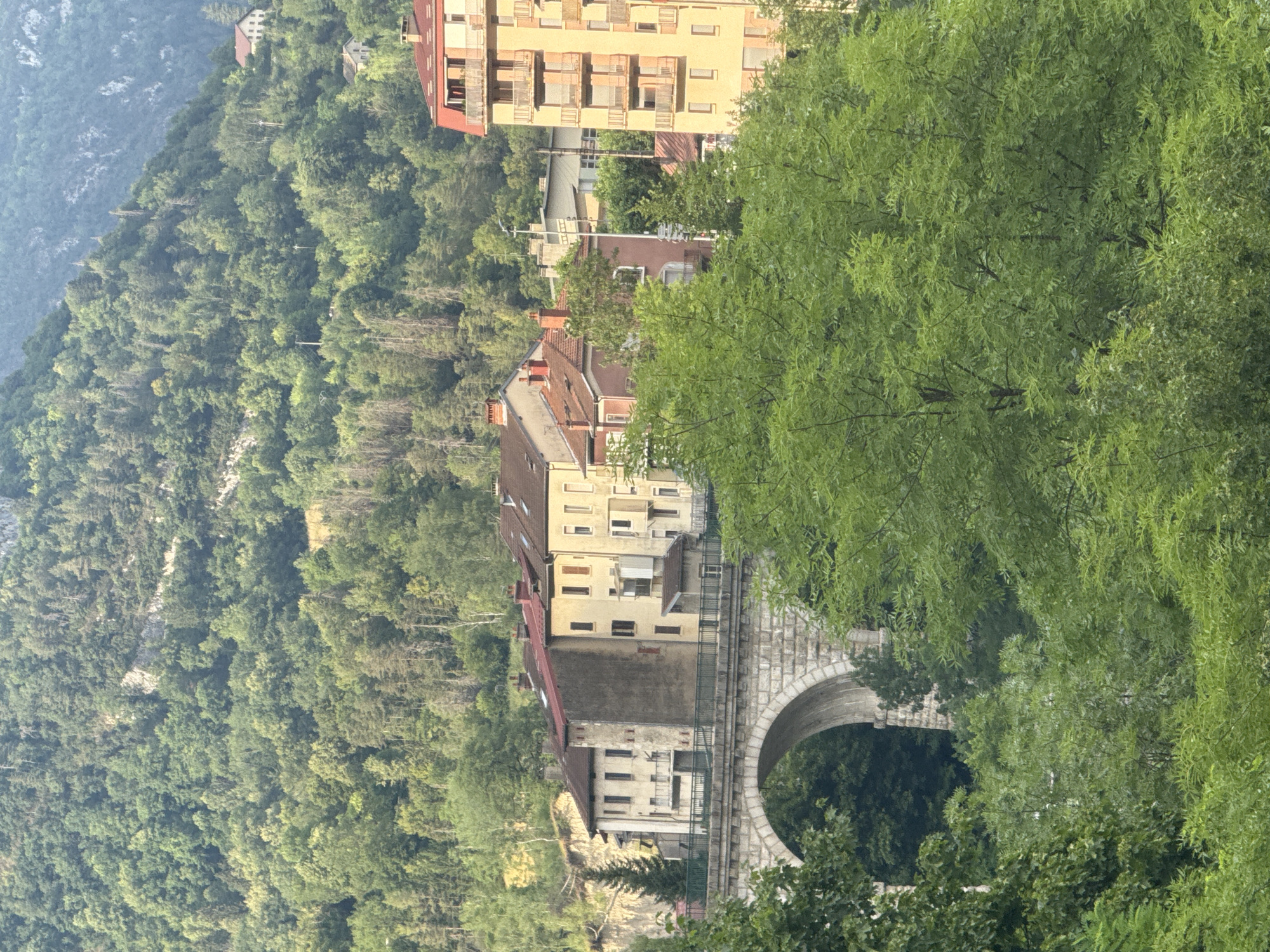 The image depicts a village nestled on a steep, tree-covered mountainside in Saint-Claude, France. Several multi-story buildings, characterized by their yellowish-beige facades and red-tiled roofs, are visible along the slope. Some buildings have balconies, and many feature numerous windows. A stone bridge with a large arched opening is a prominent feature, suggesting it spans a gap or waterway below. Lush green trees, with dense foliage, dominate the foreground and the surrounding landscape, creating a natural and verdant setting. The time of day appears to be late afternoon or early evening, given the soft lighting and the slight shadows. The weather seems pleasant, with no indication of rain or harsh sunlight. The overall scene conveys a sense of tranquility and a harmonious blend of human habitation with the natural environment. There is no visible text in the image.