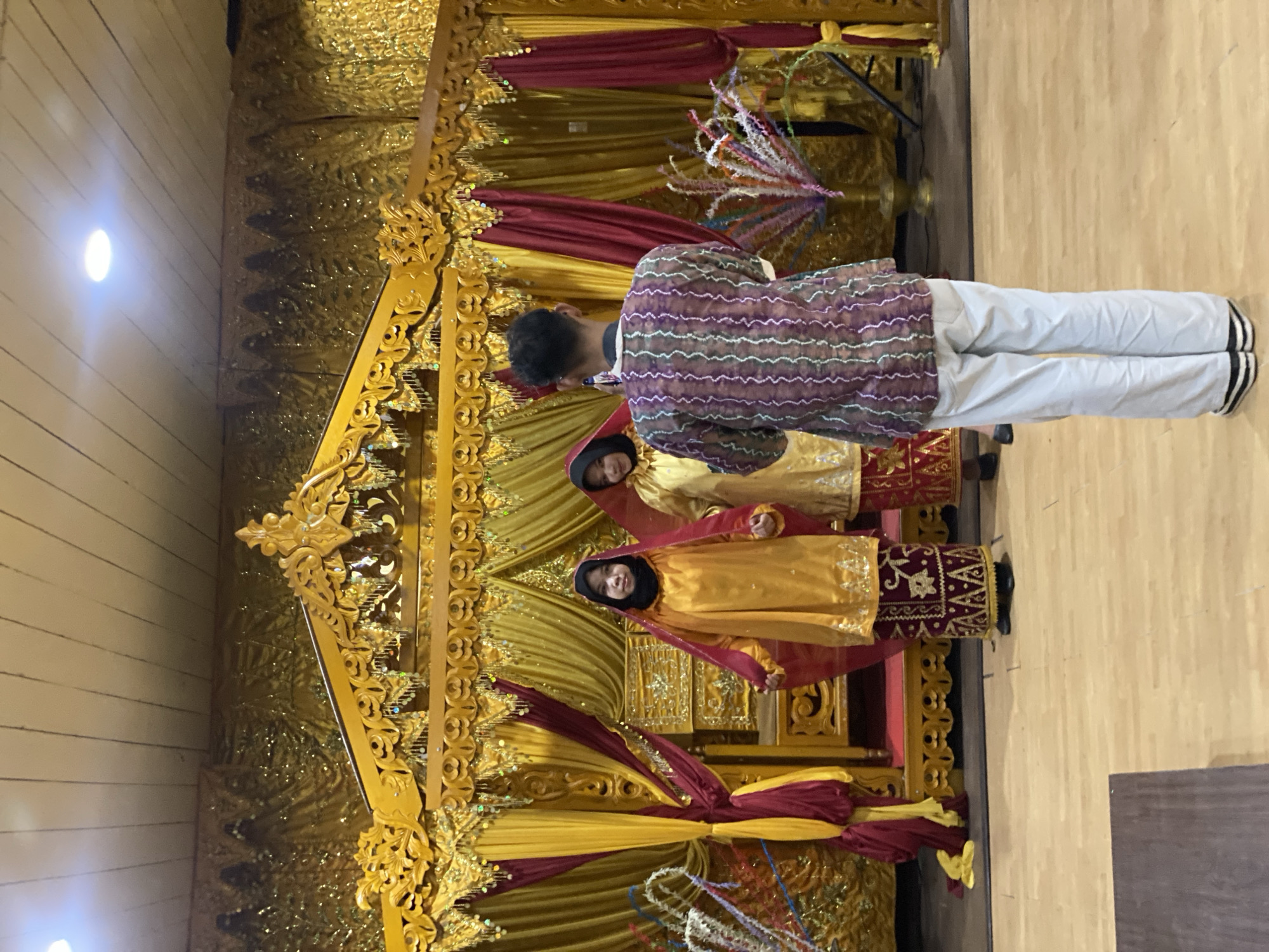 A man, seen from the back, stands on a wooden floor in front of a stage. He wears a patterned shirt and light-colored pants. On the stage, two women in traditional attire, one in yellow and red, the other in gold and red, stand facing the audience. The stage is adorned with gold and red drapes and intricate wooden carvings. The scene appears to be a cultural event, possibly a wedding or performance, set in Banjarmasin, Indonesia.