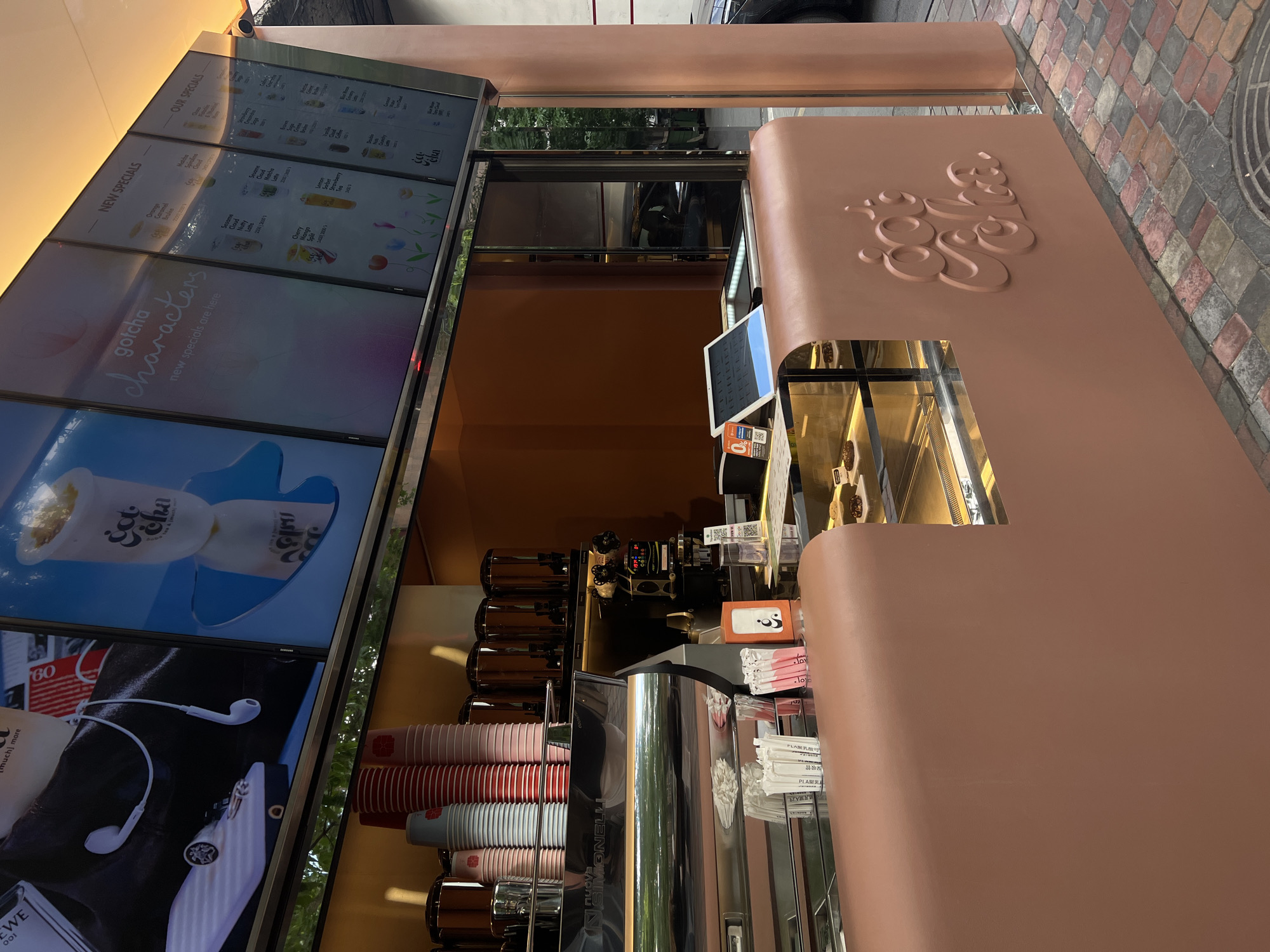 An eye-level shot captures a modern beverage kiosk, likely a bubble tea or coffee shop, situated outdoors on a paved area in Yerevan, Armenia. The kiosk features a distinctive peach-colored counter with a raised, stylized "gotcha" logo on the front. Behind the counter, various machines and stacked cups are visible, including what appears to be a Nuova Simonelli espresso machine.
The kiosk is equipped with several vertical digital display screens showcasing its menu and promotional content. Text visible on these screens includes "NEW SPECIALS," "OUR SPECIALS," "gotcha characters," and "new specials are here." Specific drink names like "Orange Caramel Bruline," "Matcha Sesame Cloud," "Mango Cloud," "Lemon Strawberry," and "Cherry Mango Splash" are displayed, alongside what seems to be "Famos Nutty Latte." One screen also shows an image of headphones and a partially visible "Loewe" logo on a bag.
Behind the counter, various colorful cups are neatly stacked, including red, white, pink, and gray. Small signs with "PLA 深吸管" (PLA deep straw) are also seen. The background shows reflections in glass, suggesting a building or trees opposite the kiosk, and textured brick-like pavement on the ground. The lighting indicates it is daytime.