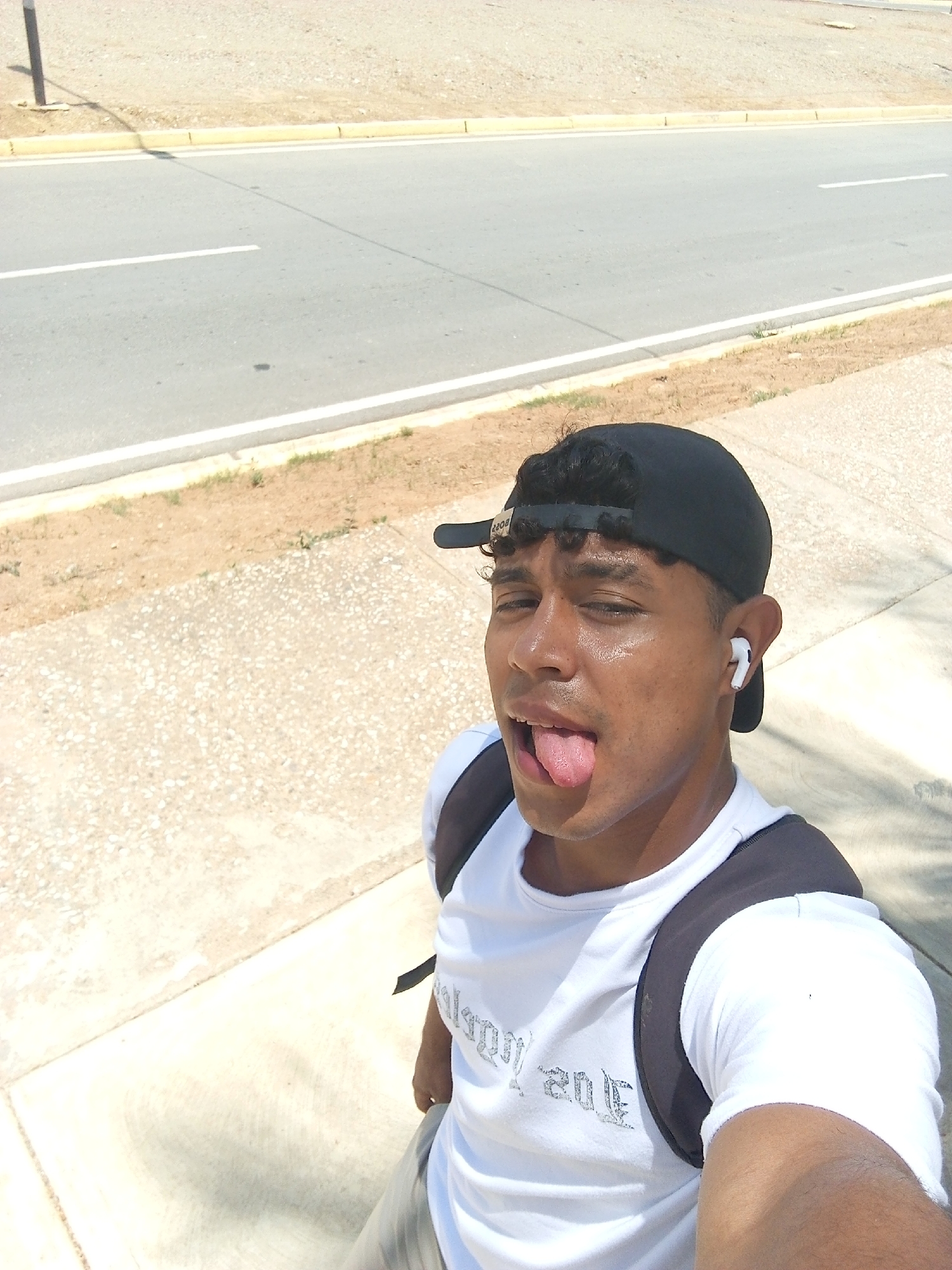 A young man is captured in a sunlit outdoor selfie, likely on a street in Porlamar, Venezuela. He is positioned on a light-colored paved sidewalk, with a two-lane asphalt road featuring white lane markings and a yellow curb behind him. Beyond the curb is a dry, sandy, or dirt shoulder. The bright sunshine suggests it is daytime, possibly mid-day, and his glistening face indicates warm or hot weather.
The man, who has tan skin and short, curly dark hair visible under his backward-worn black baseball cap, is looking directly at the camera. He is making a playful expression, with his tongue extended. He is wearing a white t-shirt with stylized, possibly Gothic-style text that appears to read "Los Angeles" across the chest. A dark-colored backpack is visible on his shoulders. A white wireless earbud is present in his right ear. In the distant background, a thin pole casts a distinct shadow on the road. The overall scene depicts a casual, bright moment on a typical street in a warm climate.