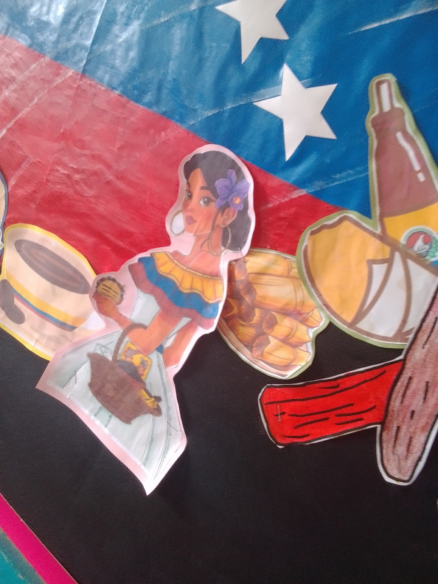 This media file is a photograph of a colorful display, likely a bulletin board or a wall decoration, featuring cutout figures and drawings. The background appears to be a large fabric or paper with the colors of the Venezuelan flag - yellow, blue, and red stripes with white stars on the blue stripe.
The main subject is a cutout figure of a young woman drawn in a cartoon style. She has dark hair adorned with a purple flower and is wearing a traditional-looking dress with yellow, blue, and red ruffles around the neckline. She is holding a round object, possibly food like a arepa or a hamburger, in her right hand and a woven basket in her left. The basket contains some yellow objects that might be fruit or vegetables.
Other cutout figures and drawings are present. To the left of the woman is a representation of a drum with yellow, blue, and red accents. To her right, there is an image of several tamales, a traditional Venezuelan dish, wrapped in corn husks. Further to the right, a brown bottle with a label is depicted, and next to it, a drawing of what appears to be a piece of meat with a red strip across it, resembling a cut of beef.
The overall impression is a festive and culturally themed display, possibly related to Venezuelan traditions, food, or celebrations. The lighting suggests it might be daytime, and there are no clear indications of weather or a specific time of day. The bright colors and cheerful style of the cutouts evoke a sense of joy and cultural pride. No visible text is present in the image. The context provided indicates the location as La Tagua, Venezuela.