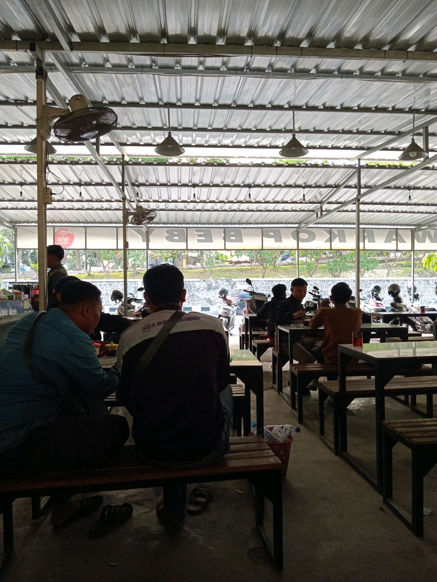The image depicts the interior of an open-air restaurant or warung in Randuagung, Indonesia. The structure has a corrugated metal roof supported by metal beams and poles. Hanging from the ceiling are industrial-style ceiling fans and pendant light fixtures. Several tables and benches are arranged in rows on a concrete floor.
In the foreground, two men are seated at a table, seen from behind. The man on the left wears a blue shirt, and the man on the right wears a dark jacket with white and purple accents and text. Both have bags slung over their shoulders. Another man, wearing a dark uniform or jacket, stands behind the man in the blue shirt.
In the middle ground, several more tables and benches are visible, with other patrons seated at them. To the right of the tables, a red trash can filled with plastic bottles is positioned near a table.
In the background, through a large window or opening, a street scene is visible. Motorcycles are parked in front of the establishment, and trees and greenery line the background. A large sign is mounted on the exterior of the building, with some of the letters visible reading "WARUNG". The overall atmosphere suggests a casual dining setting.