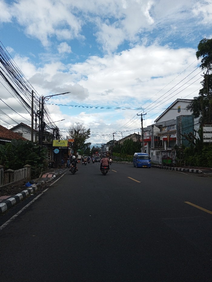 This image depicts a street scene in what is identified as the city of Serpang Shalli, Bhutan, under a partly cloudy sky. The asphalt road, appearing dry, extends from the foreground into the distance, with two solid yellow lane markers visible on the right side. Black and white striped curbs line both sides of the roadway.
On the left side of the road, a low concrete wall is partially obscured by lush green foliage. Behind this, a building with a red-tiled roof is visible. Further along the left, another building displays a sign reading "H.M. BASKA" and a circular blue sign below it; several indistinct individuals are present near this storefront. A dense network of black utility wires is strung between multiple utility poles, one of which supports a black streetlamp extending over the road.
On the right side of the road, similar black and white striped curbs are present. A row of trees and green shrubs separates the road from multi-story buildings. A prominent light-colored building with a red-tiled roof and red accents is visible, followed by another lighter building featuring a red and white emblem. Overhead, additional utility wires are visible, though less dense than on the left. A blue minivan is positioned on the far right side of the road, facing away from the viewer.
Traffic consists primarily of motorcycles, all traveling away from the viewer. The closest motorcycle on the left is black, ridden by a helmeted individual. Another motorcycle in the center is ridden by a person wearing a light-colored shirt and helmet. Multiple additional motorcycles are visible further down the road, indicating moderate traffic flow. In the background, distant hills or mountains are faintly discernible beneath the white cumulus clouds scattered across the blue sky.