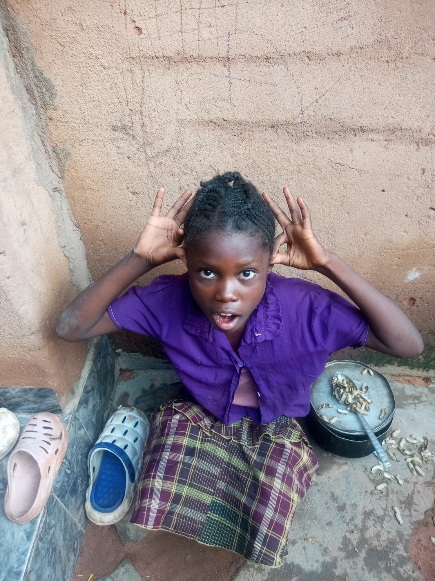 In Tempure, Nigeria, a young girl with braided hair, wearing a purple shirt and a plaid skirt, sits on a concrete floor leaning against a textured, brown wall. Her hands are raised to her ears, and her mouth is open as if she is speaking or expressing surprise. Next to her is a metal pot containing a spoon and what appears to be groundnuts. Several groundnuts are scattered on the floor around the pot. To her left, on a raised concrete ledge, are two pairs of casual footwear: a light pink clog and a blue and white sandal. The scene suggests an outdoor setting, likely a domestic environment in a Nigerian village.