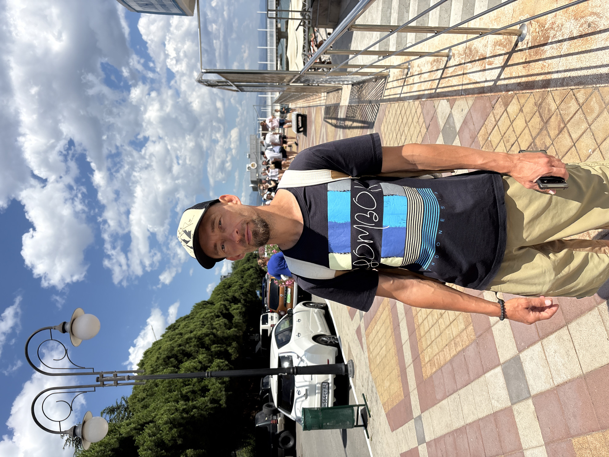 The image shows a man standing on a sidewalk in the city of Alushta, Ukraine. He is wearing a black t-shirt with a colorful striped design and the word "BOMBON" printed on it, khaki shorts, a baseball cap, and a backpack. He is holding a phone in his right hand. The background shows a street with cars parked on the side, trees, a lamppost, a railing, and a body of water in the distance with people walking along a promenade. The sky is blue with white clouds.