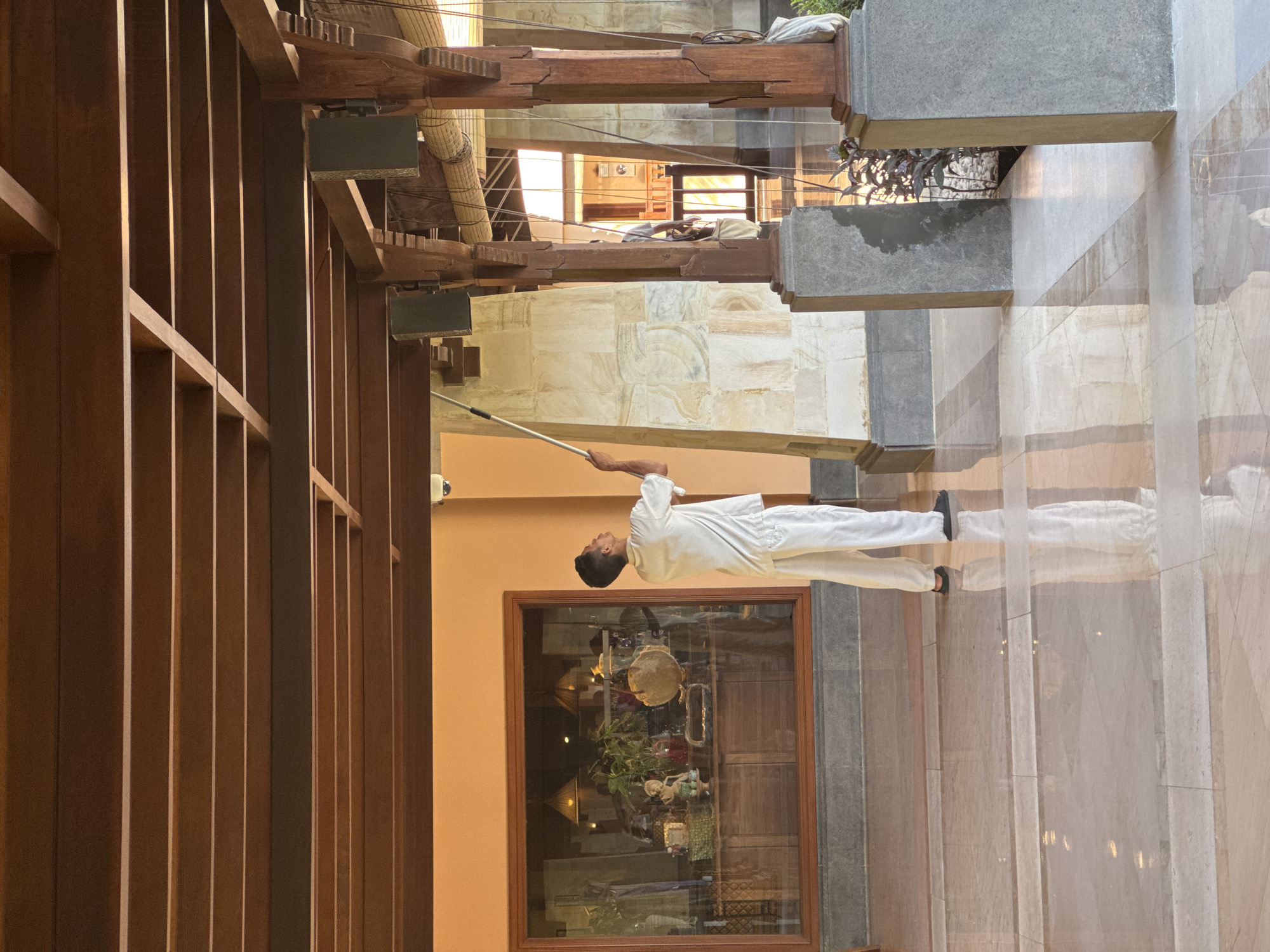 A man in white uniform holds a long pole in a corridor with wooden beams and columns in Kuta Selatan, Indonesia. The floor is light-colored marble. A large window with a wooden frame is visible behind him, reflecting interior objects. The corridor is open to the outside with some plants visible beyond the beams and columns.
