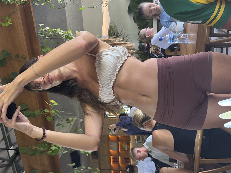 In an indoor establishment located in Tibubeneng, Indonesia, a young adult female is centrally positioned, raising both arms overhead while holding a black object, likely a smartphone, as if taking a photo. She possesses long, dark hair with bangs and wears a light-colored, ruffled crop top that exposes her midriff, paired with dark maroon tight shorts. A thin bracelet is visible on her left wrist, and a ring is on her left middle finger.
The environment features a wooden ceiling structure from which green leafy vines trail. In the blurred background, several individuals are seated at wooden tables. To the left, a male in a white shirt and a female in a black tank top are partially visible. Further back, another male wears a dark blue shirt with sunglasses perched on his head. To the right, two females are seated at a table where a plastic water bottle and a white mug are present. Part of a person wearing a green and yellow garment is visible in the immediate foreground on the right.