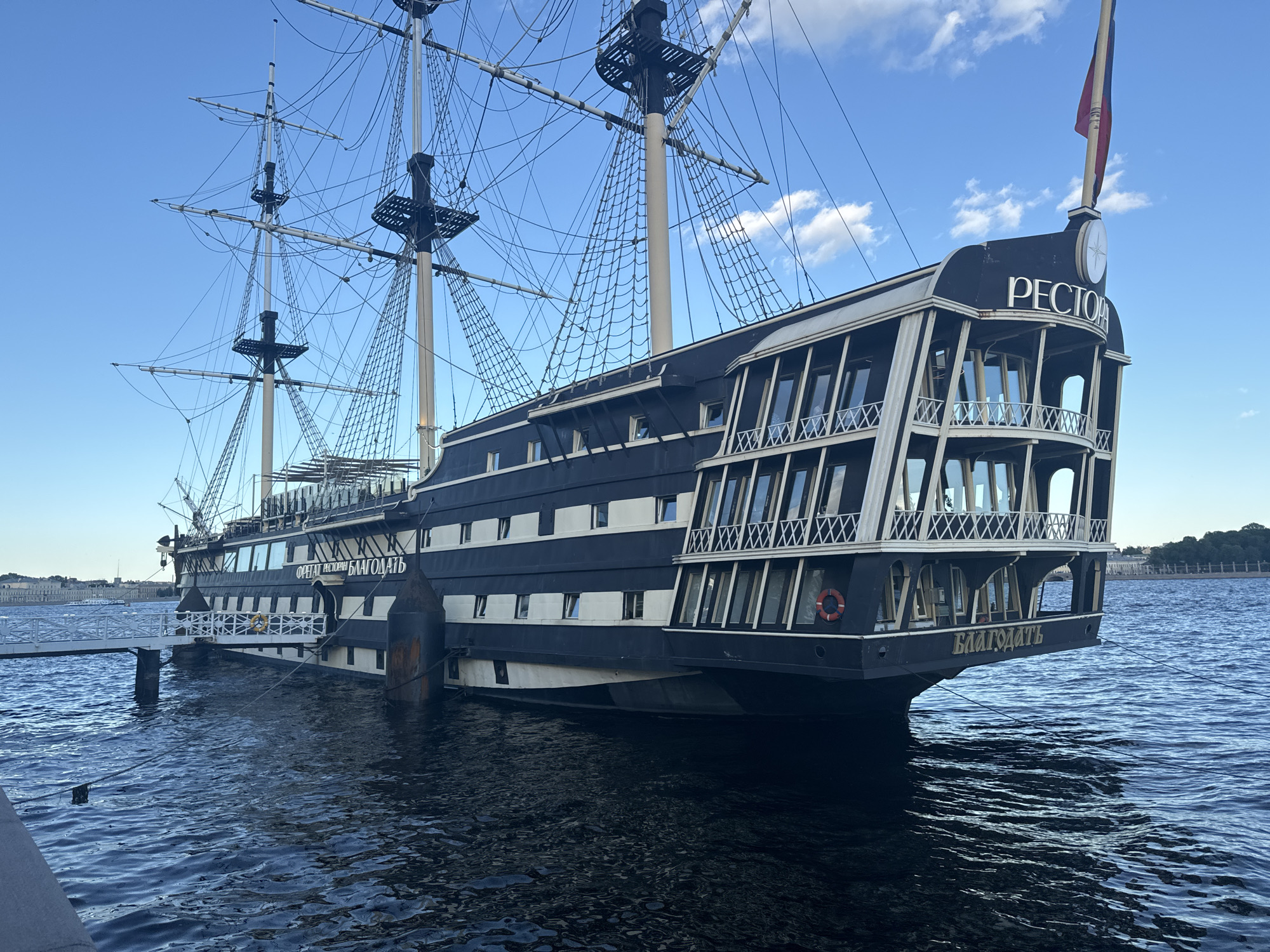 This is a medium-shot photograph of a docked, replica tall ship with the name "Благодать" (Blagodat') visible on its stern. The ship is painted black with white trim and features multiple decks with large windows. Rigging and masts are prominent above the ship. The ship is situated on dark blue water with small waves. A white gangway connects the ship to a pier on the left side of the frame. In the background, across the water, a city skyline with buildings and trees is visible under a bright blue sky with scattered clouds. The scene appears to be in Saint Petersburg, Russia, given the architectural style of the background and the presence of a historical tall ship. The lighting suggests it is daytime. There are no people visible in the image.