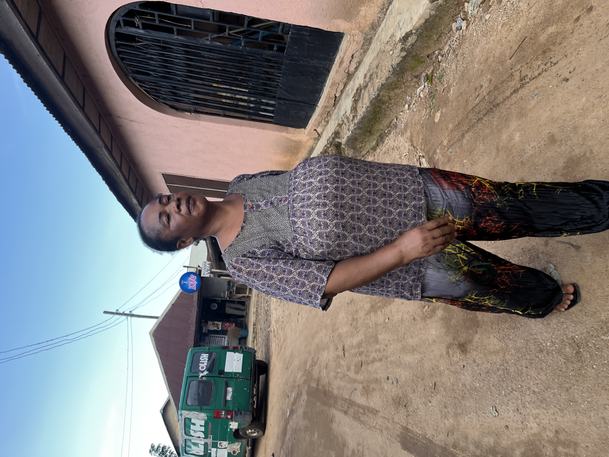 A medium shot depicts a woman standing in a dirt road, with a pink building with an arched metal gate to her right. The woman is facing slightly to her left, with her eyes closed and her head tilted upward. She has dark skin and short, dark hair. She is wearing a patterned blue and white blouse with a gray and white patterned top and black pants with a colorful, abstract design. Her feet are clad in black flip-flops.
Behind her, a green van is parked on the left side of the road. The van has "KLISH" printed on its side in white letters. Further down the road, there is a small building with a corrugated metal roof and a blue sign that says "Life." There are also some people visible in the background, standing near the small building. The road is unpaved and dusty, with some debris scattered on the ground. The sky is overcast. This scene is located in Rafin Par, Nigeria.