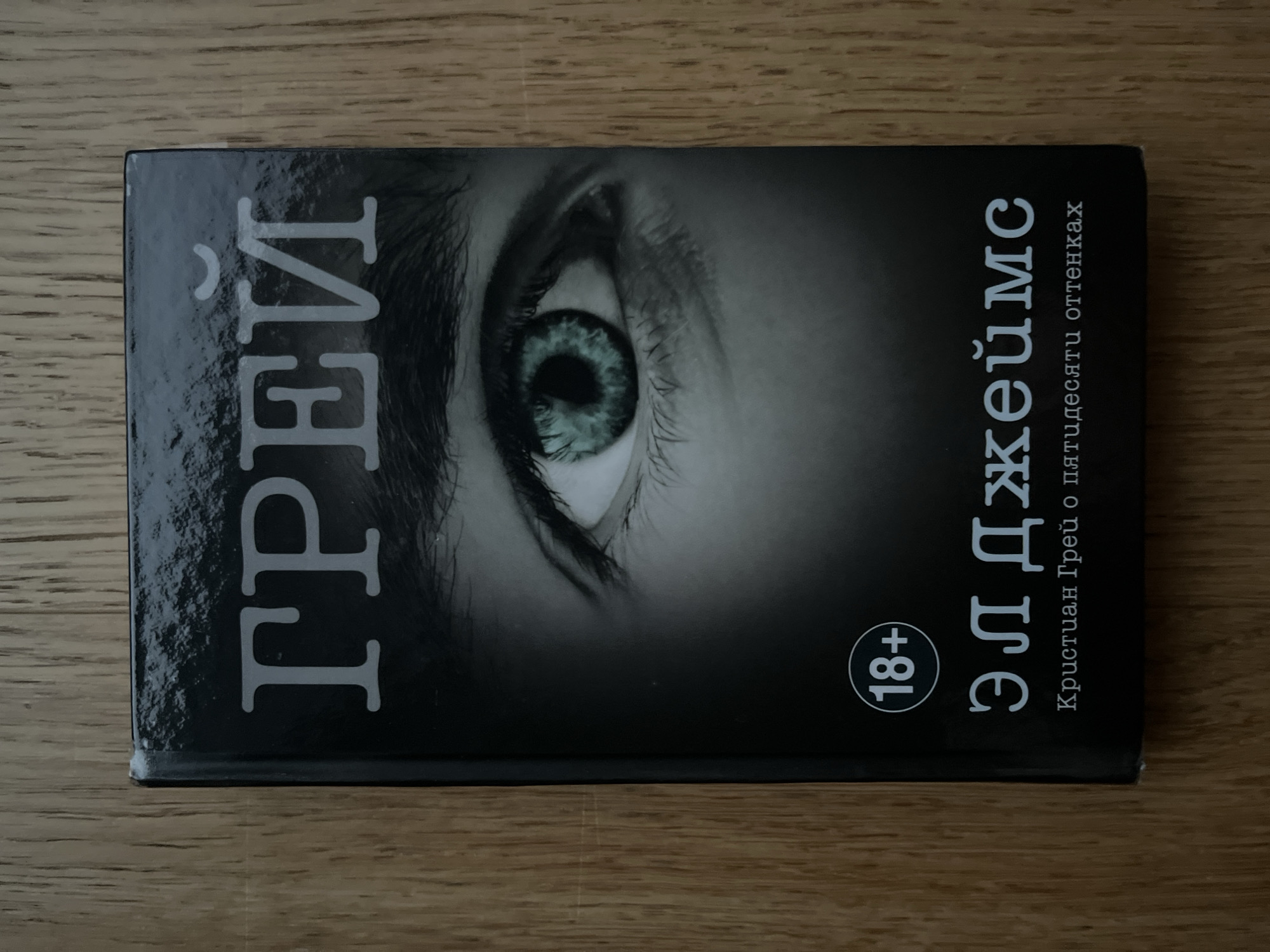 A book with a black cover is lying on a wooden surface. The cover features a close-up of a person's eye with a green iris and dark eyelashes. The title "ГРЕЙ" is printed in large, light-colored letters at the top of the cover. Below the eye image, there is a circular icon with "18+" inside. Further down, the name "ЭЛ ДЖЕЙМС" is written in large letters, followed by smaller text that reads "Кристиан Грей о пятидесяти оттенках". The book appears to be a Russian edition of "Grey" by E.L. James. The wooden surface appears to be a table or countertop. There are no people or actions depicted in the image. The location of Haabneeme alevik, Estonia is not visible or suggested in the image.