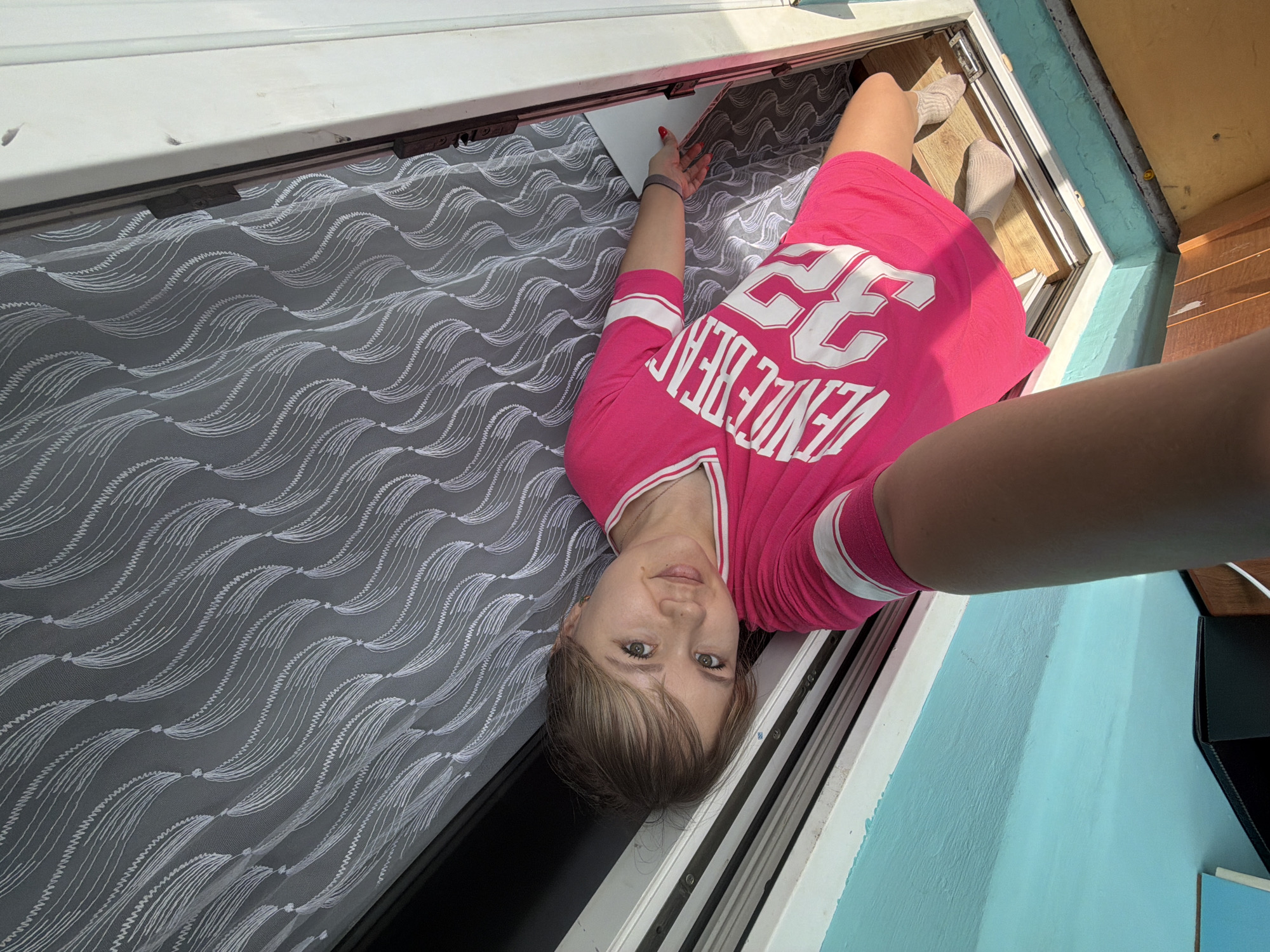 A woman lies on a pull-out bed located on a balcony, presumably in a residential setting in the city of Chaikovsky, Russia. The woman wears a pink jersey with white stripes on the sleeves and "Venice Beach 22" printed on the front. She is holding a piece of paper in her left hand. The bed is covered with a grey patterned textile. The balcony has a turquoise wall.