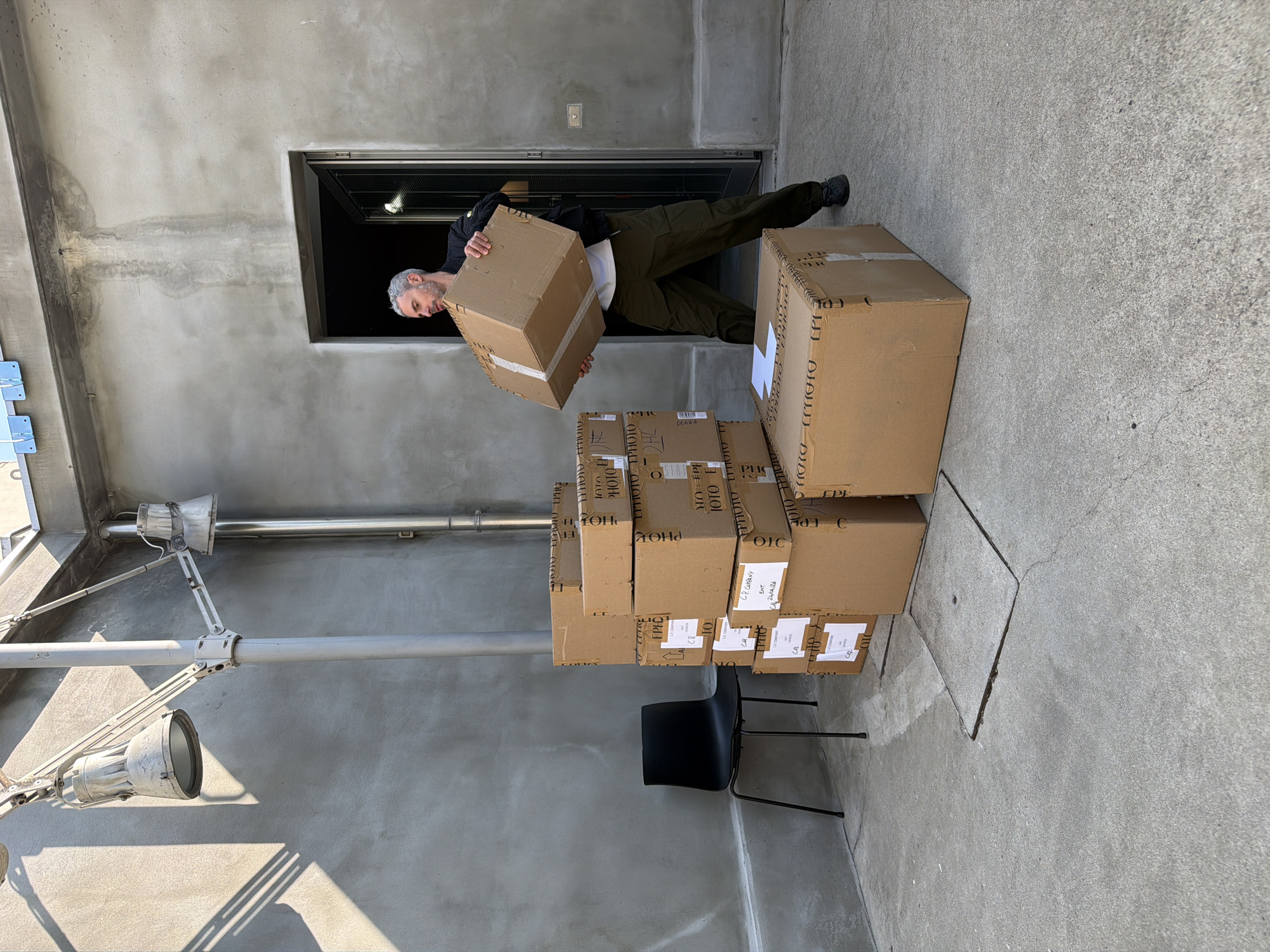 An upright shot captures a man engaged in handling cardboard boxes in an industrial or utility area, possibly in Milan, Italy. The scene is dominated by concrete surfaces under bright daylight, casting crisp shadows.
The central subject is a man with grey hair and a short beard, wearing a dark jacket over a white shirt and olive green cargo pants. He is seen emerging from a dark, rectangular doorway, holding a medium-sized cardboard box with both hands, appearing to be in the process of carrying or moving it. His expression is focused and neutral.
Around him, numerous brown cardboard boxes are stacked on the concrete ground. One large stack is visible to the right of the man, and several smaller boxes are piled in front of him. A black, lightweight plastic chair is positioned next to a stack of boxes. Industrial-style light fixtures with visible wiring and pipes are mounted on the concrete wall to the left, also casting shadows. The concrete wall shows distinct water stains or weathering marks. The ground features a visible square manhole cover.
Visible text on the boxes includes partial words and codes such as "EPHD", "PHOTO", "C8", "HOH", and notably, "CP. COMPANY" along with a date "24/04/26". These details suggest the boxes contain merchandise, possibly related to the CP Company brand, and indicate a shipping or delivery operation. The bright, direct sunlight suggests it is daytime.
