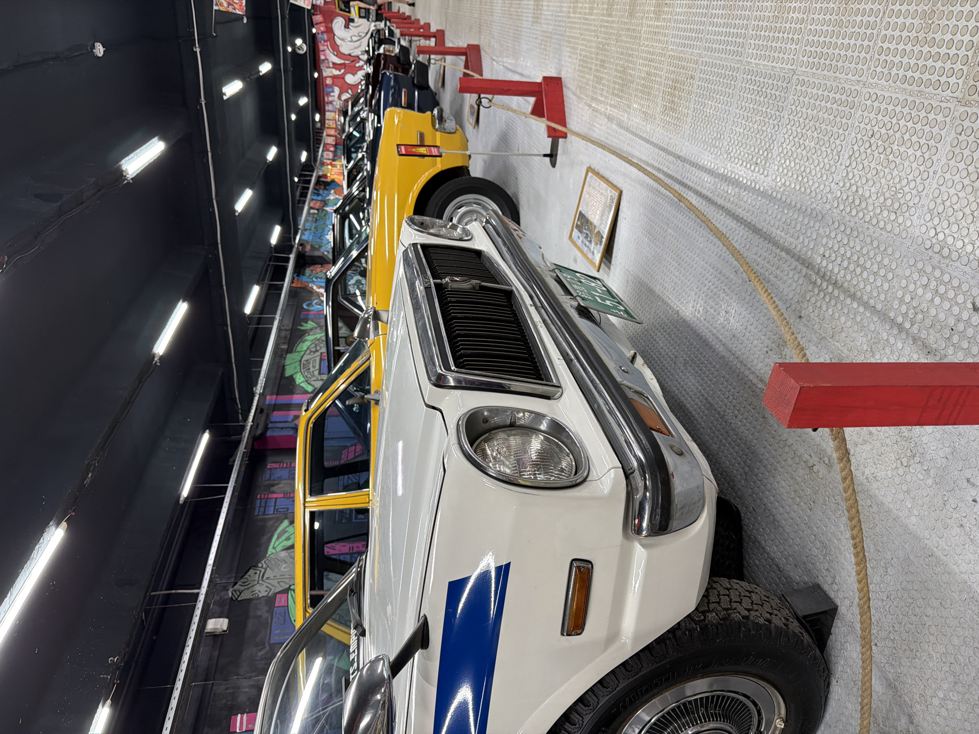 In Moscow, Russia, a row of vintage cars is displayed indoors, likely in a museum or exhibition space. The foreground features the front of a white car with a blue stripe, its headlight and grille prominently visible. Behind it, a bright yellow sedan is parked, followed by several other sedans in darker colors, including navy and black, receding into the distance. The cars are lined up neatly, separated by red stanchions and a thick rope barrier. The walls are decorated with colorful graffiti art, depicting stylized cityscapes and abstract designs, illuminated by fluorescent lights on the black ceiling. The floor is a textured, light-colored surface with a repeating circular pattern. A framed document is placed on the floor in front of the yellow car. No people are clearly visible in the image.