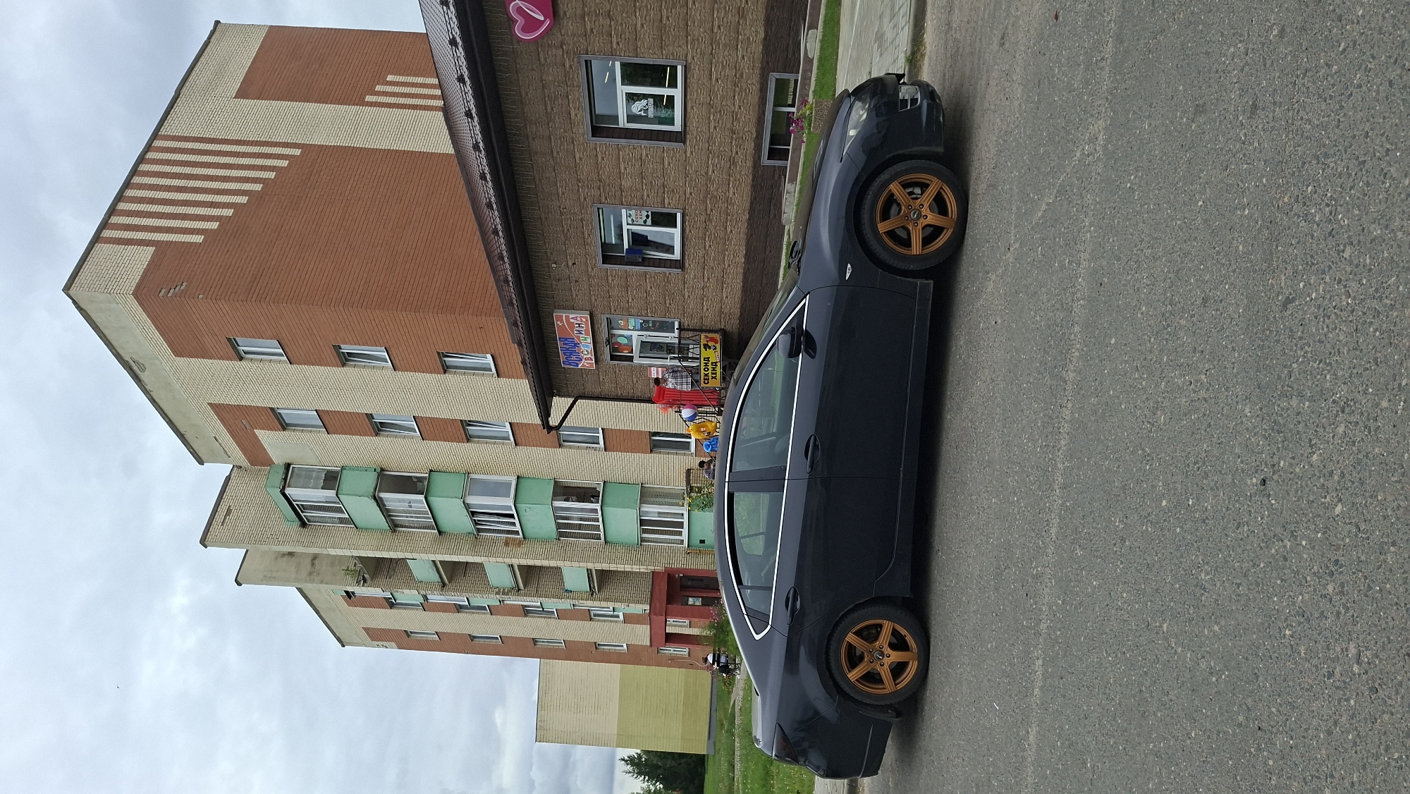 A dark blue sedan with bronze rims is parked on the side of a paved road in front of a brick building. The building has a brown, textured facade and a dark brown roof. There are several windows on the building, two of which are visible and rectangular with white frames. Above the windows, a sign with red lettering and a pink heart graphic is visible. To the left of the brick building is a larger, multi-story apartment building with light beige and orange brickwork. This apartment building features several balconies with green trim. The sky is overcast and cloudy. In the background, a lighter colored building and some trees are visible. The scene appears to be a street in the city of Myadzyel, Belarus, likely in a residential or commercial area.