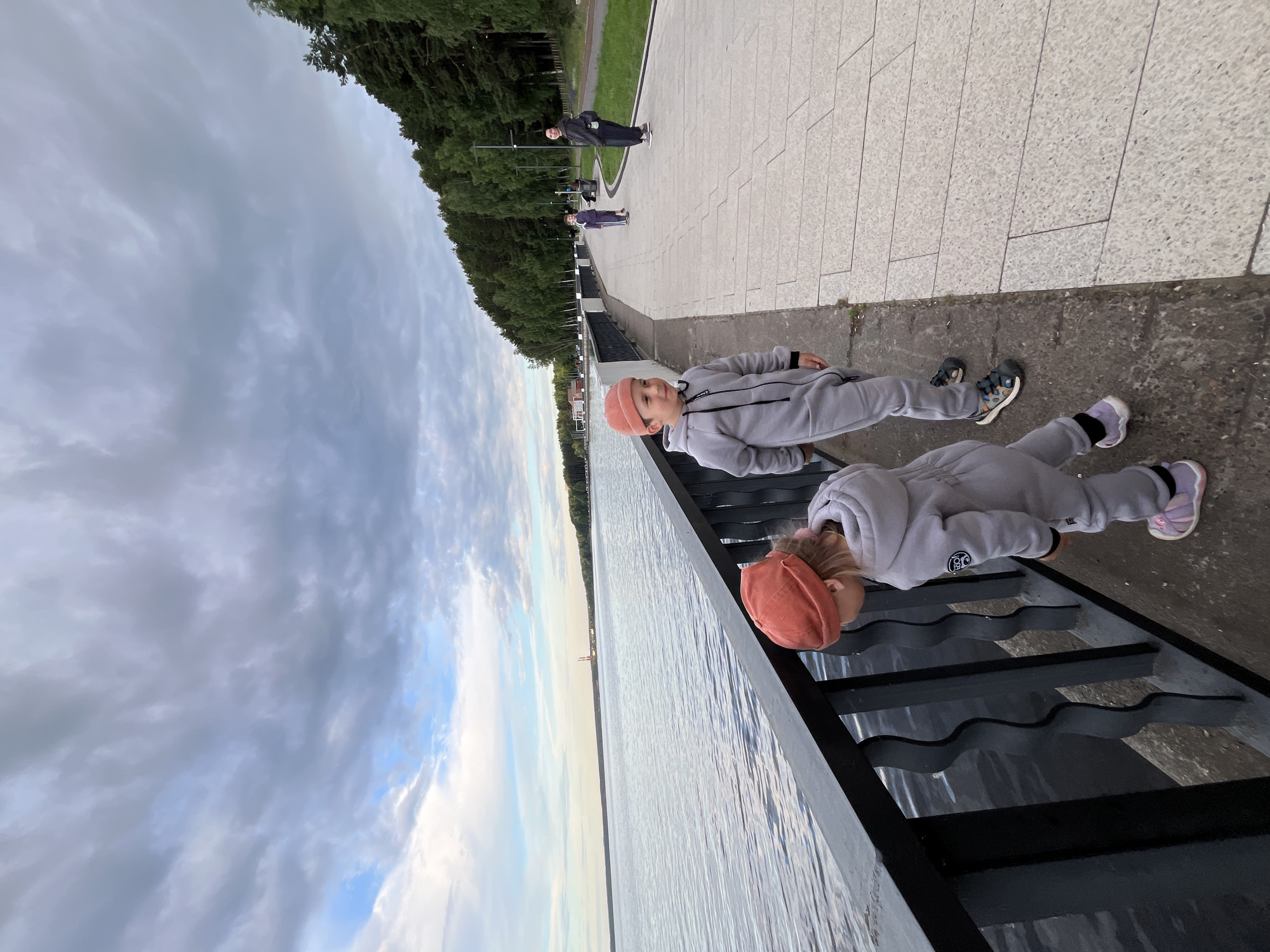 In Chaikovsky, Russia, two young children are the central subjects in this outdoor photograph. They are dressed in light grey tracksuits and orange beanies, appearing to be siblings or friends. The children are positioned on dark, wavy metal stairs adjacent to a body of water. One child is looking down, while the other is looking towards the camera. In the background, a paved walkway lined with trees stretches along the waterfront. A few other figures are visible further down the walkway, including two adults in darker clothing and another child in a purple outfit. The sky is filled with dramatic, cloudy formations, suggesting it might be late afternoon or early evening. The water appears calm, reflecting the diffused light of the sky. The scene conveys a casual, outdoor outing, possibly a walk by the river or lake. There is no visible text in the image.