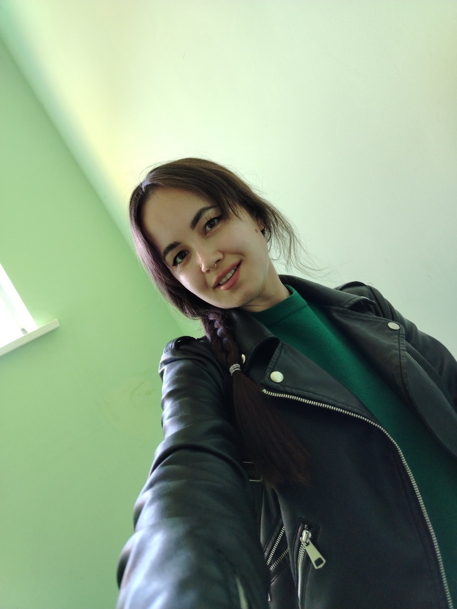 A low-angle selfie shows a young woman smiling subtly indoors, likely in Noviy, Russia. She is the primary subject, looking directly at the camera with a gentle expression. Her dark hair is styled in a braid that rests over her left shoulder. She is dressed in a dark green sweater, layered under a black leather-style jacket with silver zippers and buttons. A septum piercing is visible on her nose. The background consists of light green walls, with a white window sill visible on the left, suggesting natural daylight illuminates the room. The overall atmosphere is casual and relaxed. No text is visible in the image.