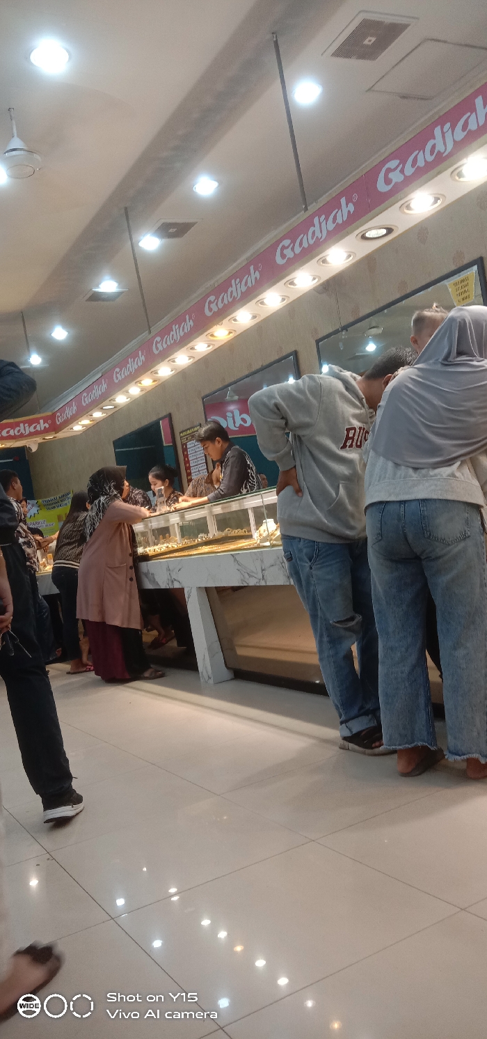 The image displays the interior of a gold retail establishment identified by prominent overhead signage reading "Gadjah," located in Probolinggo, Indonesia. Approximately ten individuals are present, predominantly customers, gathered around brightly lit, glass-topped display cases filled with gold jewelry.
The environment is a well-lit indoor space featuring a white ceiling with recessed circular lights and a visible white ceiling fan. A gray beam traverses the ceiling. The walls are light-colored, possibly textured, with a continuous pink sign bearing the white "Gadjah" logo repeated horizontally and illuminated by integrated spotlights.
Customers are observed leaning over and peering into the display cases, which have white and gray marbled bases, some with reflective lower panels. In the mid-ground, a male subject wearing a light gray hooded sweatshirt with "RUS" (likely "RUSSELL") lettering and distressed blue jeans with a torn left knee stands with hands on hips, observing the displays. Adjacent to him, a female subject wears a light gray hijab, a light gray long-sleeved top, and light blue wide-leg jeans with frayed hems. Both wear dark sandals. Further left, a woman in a light brown or peach long cardigan over a dark skirt and a dark headscarf also examines the display contents. In the background, behind the counters, staff members or additional customers are visible, one male wearing a patterned dark shirt. The floor consists of large, reflective light-colored tiles.