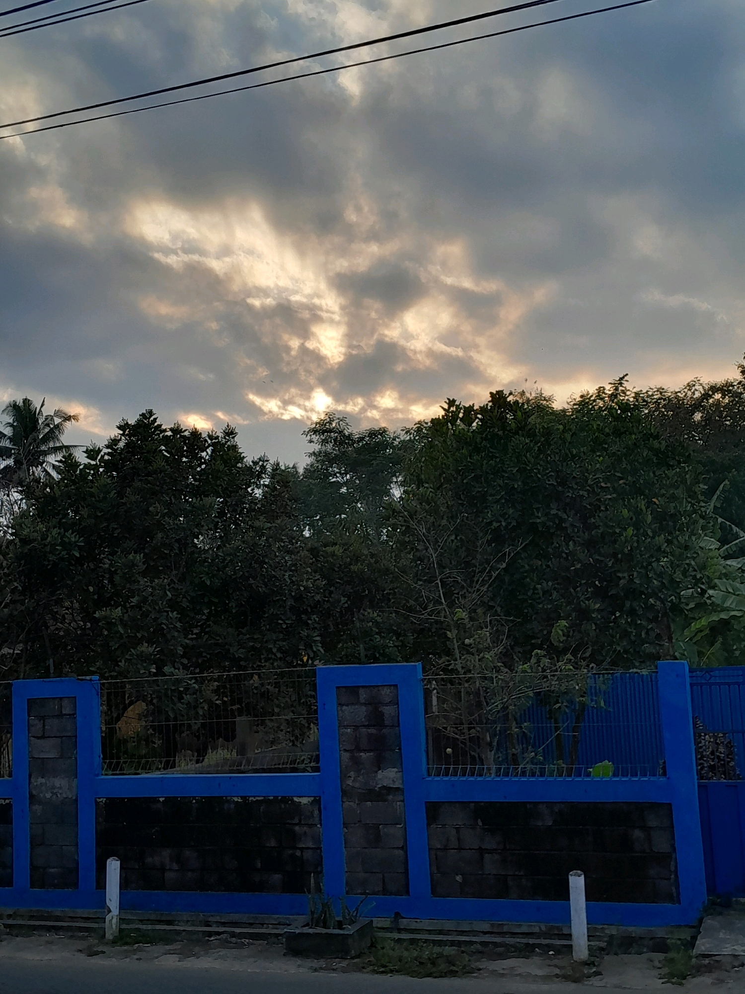 The image depicts a street view in Glanggang, Indonesia, under a cloudy sky at dusk. In the foreground, a blue-painted fence made of concrete blocks and metal bars runs horizontally across the frame. Beyond the fence, a dense line of dark green trees and foliage stretches across the mid-ground. In the background, the sky is filled with dramatic clouds, with rays of sunlight piercing through, suggesting the sun is setting or has recently risen. Power lines are visible running across the top of the frame. The overall scene is naturalistic and suggests an outdoor environment in a semi-rural or residential area.