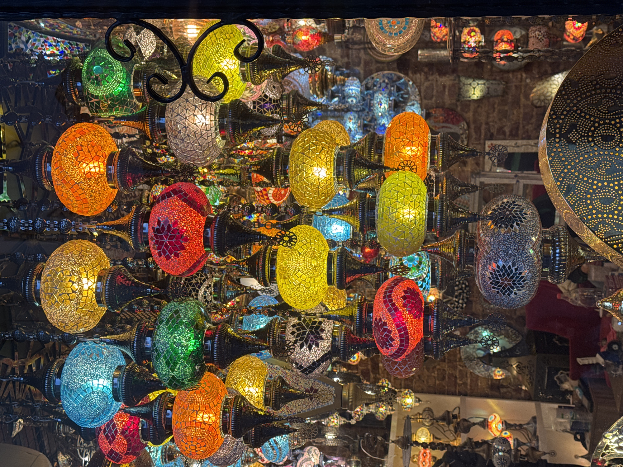 This vivid, overhead shot captures a mesmerizing display of numerous brightly lit Turkish mosaic lamps, characteristic of those found in shops and markets in Beyoğlu, Turkey. The scene is dominated by a dense cluster of these ornate lights, each crafted from countless small, faceted pieces of colored glass.
The lamps come in a rich array of colors including emerald green, fiery orange, ruby red, sunny yellow, sky blue, and clear/white, arranged in intricate patterns. When illuminated, they cast a warm, glowing light that highlights their detailed mosaic work. The lamps are supported by dark, decorative metal fixtures with elegant curves and flourishes, contributing to their traditional aesthetic. They are densely packed, creating a kaleidoscopic effect enhanced by reflections on the surfaces behind them, making the display appear even larger and more expansive.
While the main subject is the array of lamps, the reflective background subtly reveals more of the setting. In the lower right portion of the image, a faint, reddish reflection can be seen, suggesting the presence of a person or mannequin observing the display. No specific activity or event is taking place beyond the static presentation of these beautiful handcrafted items. The atmosphere conveyed is one of warmth, cultural richness, and vibrant artistry. The time of day is implied to be dark, either indoors or evening, allowing the lamps to serve as the primary source of illumination. There is no visible text in the image.