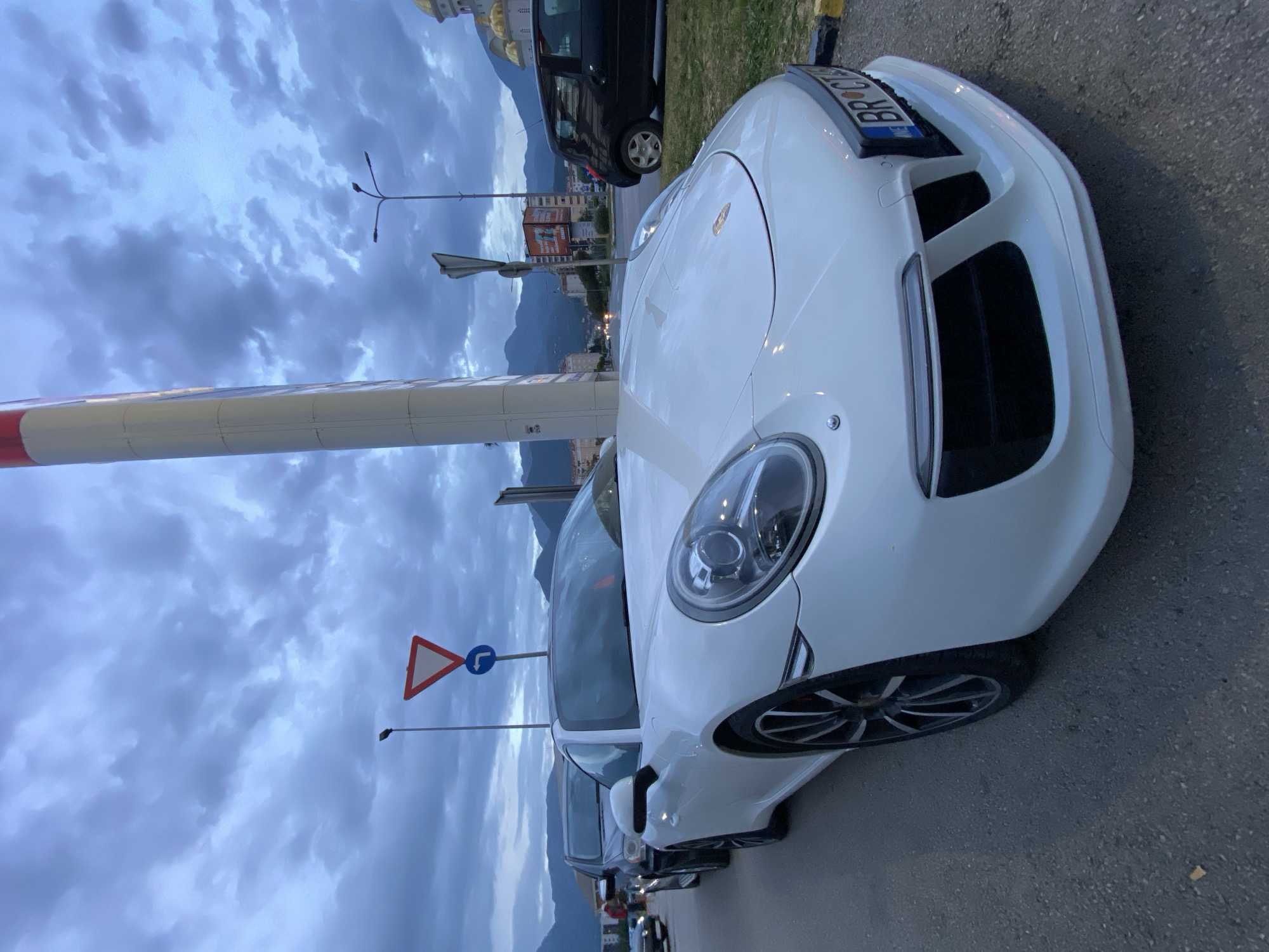 A white Porsche is parked on the side of a road in Bar, Montenegro. The vehicle is positioned near a gas station, indicated by a tall white pole with red accents. Behind the Porsche is a dark-colored minivan. In the background are mountains and buildings, including one with a golden dome, possibly a religious structure. Road signs are visible near the cars. The sky is overcast with dark clouds.