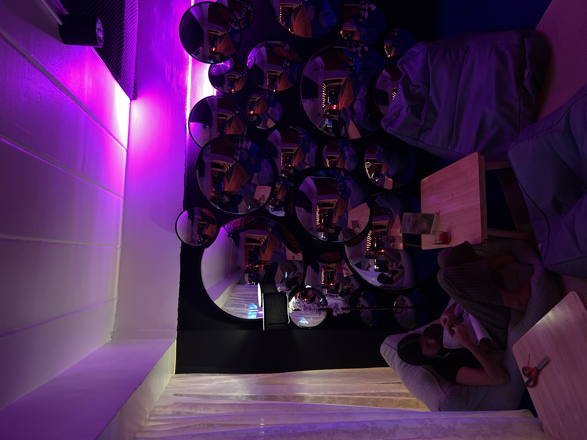 The image depicts a room in Phuket City Municipality, Thailand, featuring an artistic installation on a wall. The wall is covered with numerous round mirrors of varying sizes. The mirrors reflect various elements of the room and create a distorted perspective of the space. A woman is seated in a beanbag chair in the foreground. She is reading or sewing and there are a pair of scissors on a table next to her. Adjacent to her are additional beanbag chairs and small wooden tables, creating a comfortable lounge area. The room is illuminated with purple and white lighting. The overall atmosphere of the room is modern, with a focus on design and creating an immersive visual experience.