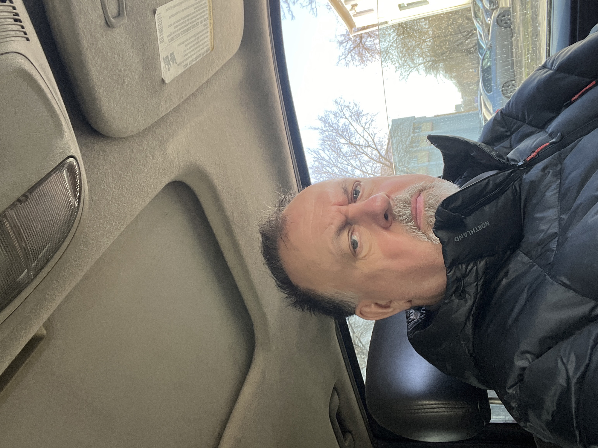 A selfie taken inside a car shows a middle-aged man wearing a black puffer jacket. The interior of the car is visible, including the roof, sunroof, sun visor, and light fixture. Outside the car window, a street scene shows buildings and trees. The location cannot be verified as Podolsk, Russia based on visual information alone.