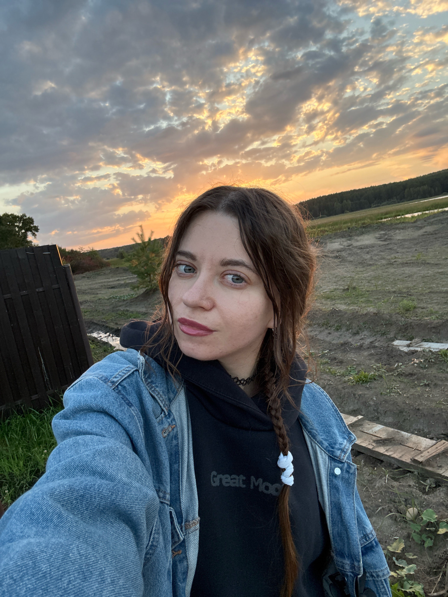 A young woman is taking a selfie outdoors at dusk. She has long, brown hair braided to one side and is wearing a denim jacket over a black hoodie. The background shows a rural landscape with a wooden fence, trees, and a body of water in the distance. The sky is filled with clouds and the setting sun, casting a warm glow over the scene. The location is identified as Pakly, Russia.