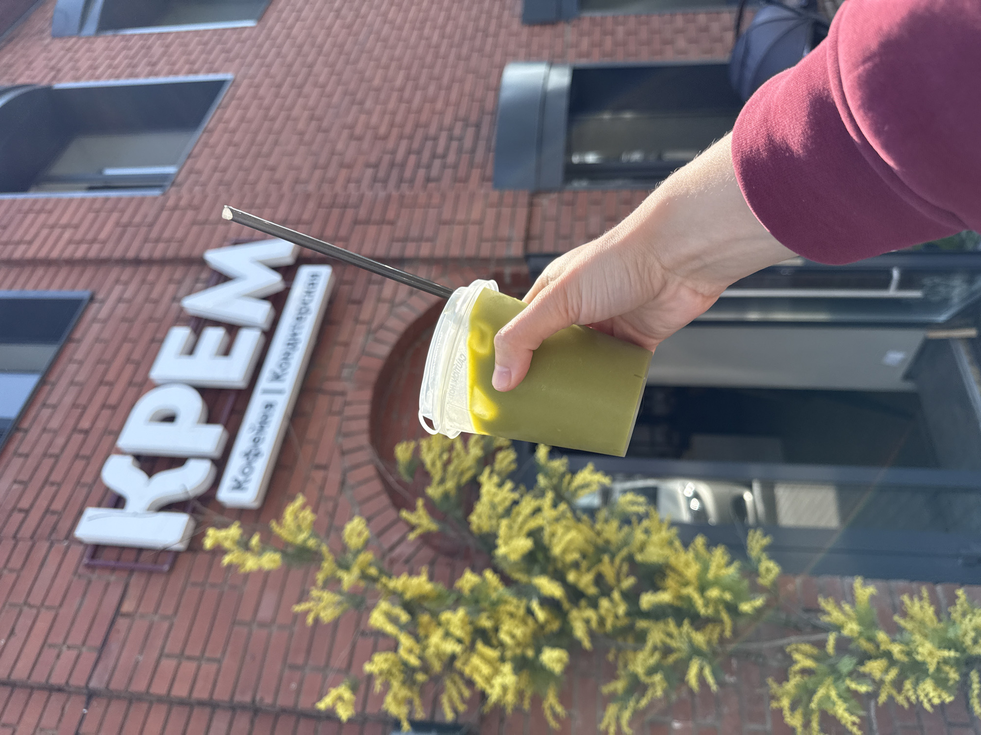 A person's hand, wearing a maroon sweatshirt, holds up a clear plastic cup filled with a vibrant green beverage, likely a smoothie or matcha drink, with a black straw inserted. The cup has a transparent lid with a slight condensation. The background features a building with a red brick facade. Above the entrance, a large, white, three-dimensional sign spells out "КРЕМ" (Krem). Below this, a smaller sign reads "Кофейня | Кондитерская" (Coffee shop | Confectionery). To the left of the building, a small tree with bright yellow foliage is visible. The overall scene appears to be a street-level view in front of a cafe or shop. The lighting suggests a sunny day. This establishment is located in Votkinsk, Russia.
