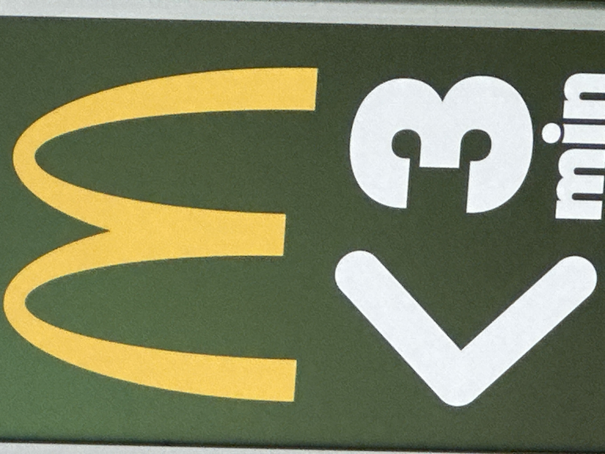 A close-up, eye-level shot captures a portion of a green sign with a large, yellow McDonald's "M" logo at the top. Below the logo, a white "<3" symbol is prominently displayed, followed by a large white number "3". Beneath the "3", the word "min" is written in white lowercase letters. The overall impression is a marketing message related to speed or time. The scene is outdoors, with the sign appearing to be free-standing or part of a larger structure. No people are visible in the frame.