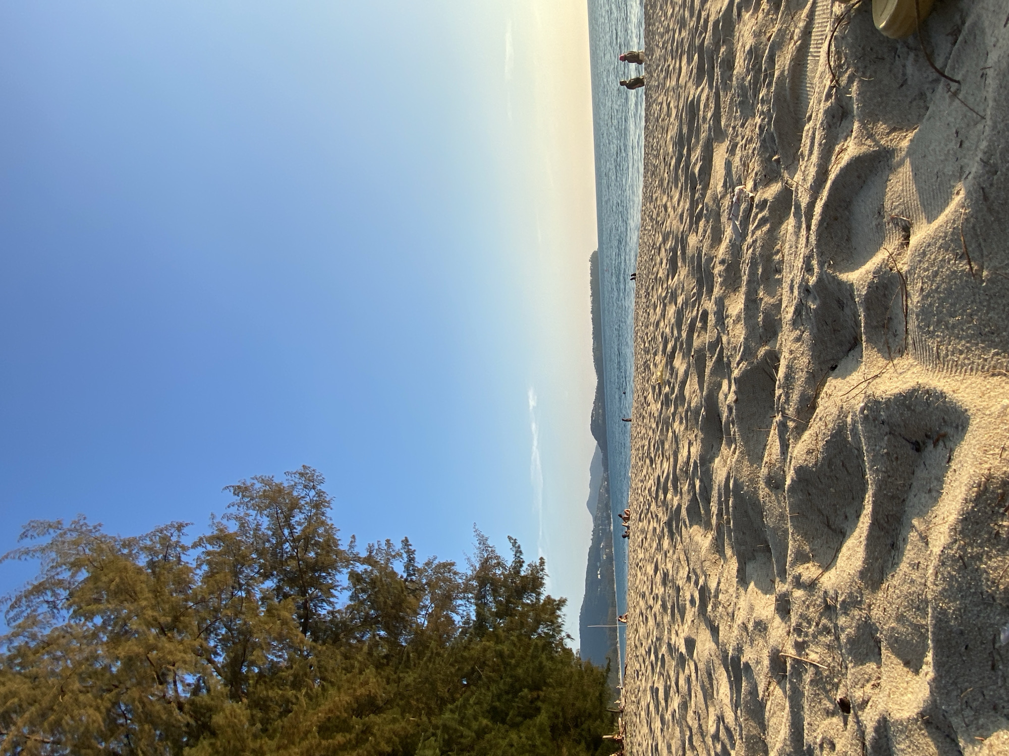 The image shows a beach scene in Choeng Thale, Thailand. The sky is blue, with a slight gradient towards the horizon. Trees are visible in the lower left corner. A sandy beach with footprints occupies a significant portion of the right side of the image. The ocean is visible in the distance. Several figures are present on the beach.