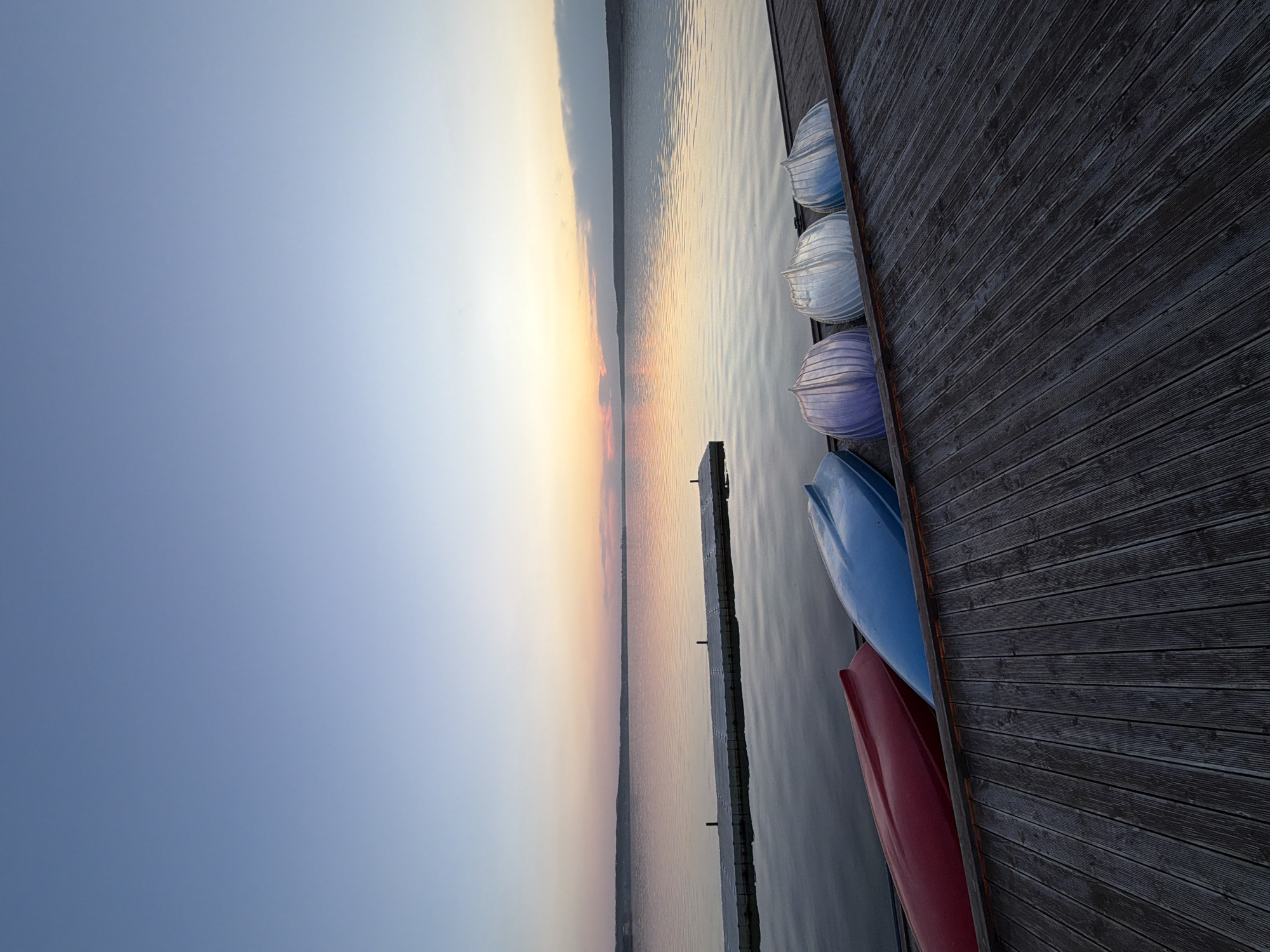 The image depicts a serene waterfront scene, likely at sunset or sunrise, featuring several canoes lined up on a wooden dock. The canoes are colored in red, blue, purple, white, and light blue. The sky is a gradient of light blue to orange, reflecting on the calm water. A thin strip of land is visible in the distance. The dock, made of dark wood, is on the right side of the image, taking up a large portion of the scene. The angle of the photo is slightly tilted. There is no evidence to confirm that this is in Votkinsk, Russia.