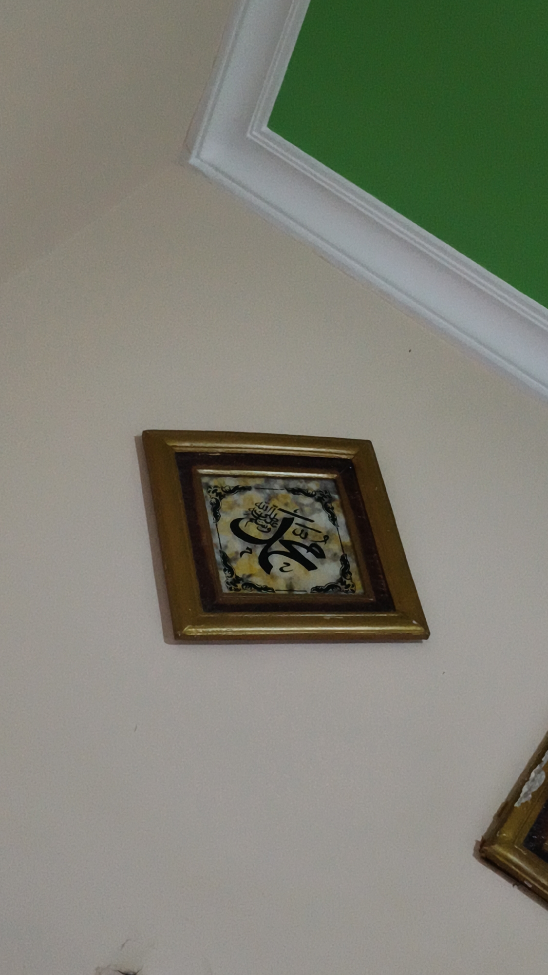 The image shows a framed Islamic calligraphy artwork hanging on a plain, light-colored wall. The artwork is square-shaped with a dark wooden frame that has a metallic gold accent. Inside the frame, the calligraphy is in black ink on a marbled background of yellow and white. Above the artwork, a white molding runs along the top of the wall, where it meets a green ceiling or upper wall section. To the right of the main artwork, a corner of another framed picture is visible, also appearing to be in a gold frame. The scene depicts an interior setting, likely a home or a place of worship, with the artwork serving as decoration. The location of Kerep Kidul, Indonesia, is not visually evident from the image itself, but is provided as context.