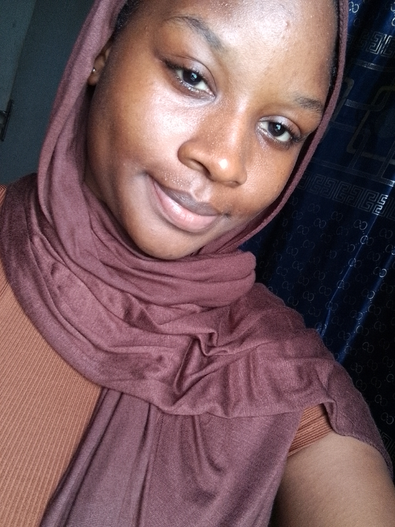 A close-up, selfie-style photograph shows a woman with brown skin wearing a brown hijab and a brown ribbed top. She is looking at the camera with a slight smile. The background features a dark blue curtain with a patterned design. The location appears to be indoors.