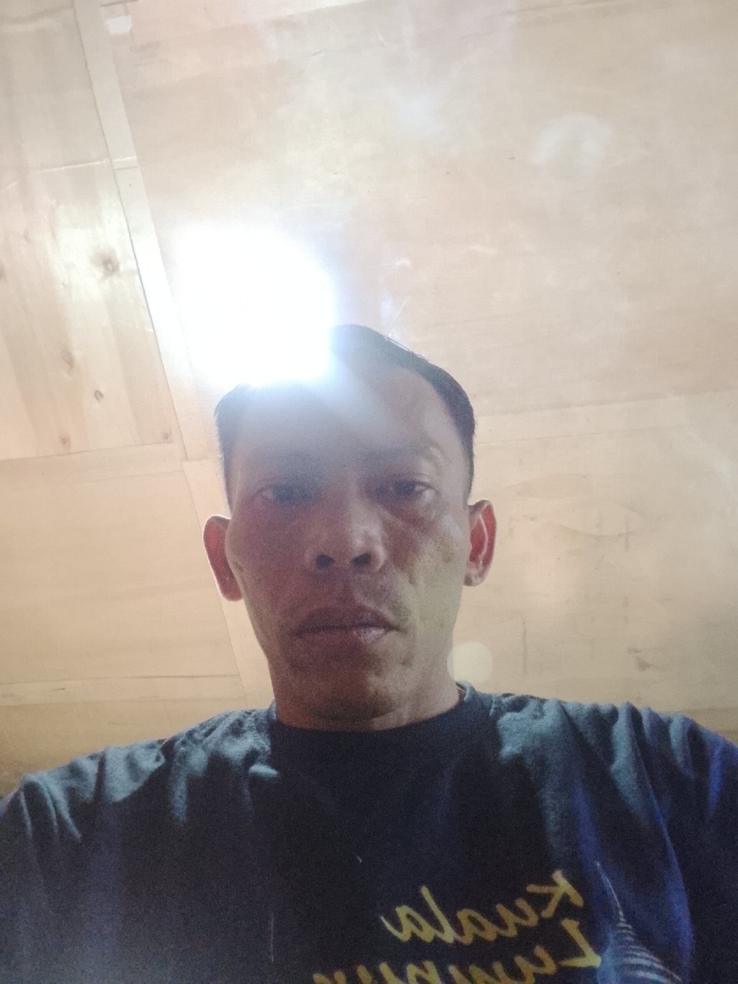 The image shows a selfie taken from a low angle, focusing on the face and upper body of a man. He has short, dark hair and appears to be indoors, with a wooden ceiling visible above him. The lighting is bright and overhead, creating a glare on the ceiling and partially obscuring the top of his head. The man is wearing a dark t-shirt with some yellow lettering on the front. His facial features are clearly visible, and he has a neutral expression. There is no indication of the location within Madukoro, Indonesia, or any specific actions or interactions taking place.