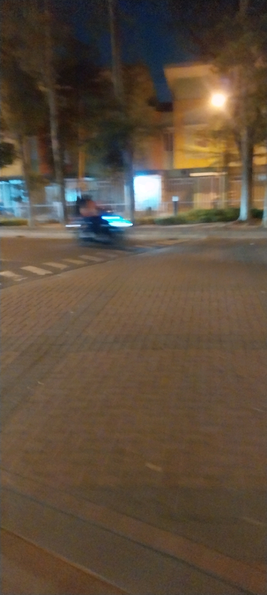 A blurry, night-time, street-level perspective captures a motorcyclist in motion, crossing a pedestrian walkway. The motorcyclist, with their form obscured by motion blur, is illuminated by the bright blue headlights of their vehicle. In the background, indistinct trees and a building with glowing windows suggest an urban environment. The ground appears to be paved with brickwork. The scene is likely in Kelapa Dua, Indonesia, given the context provided.