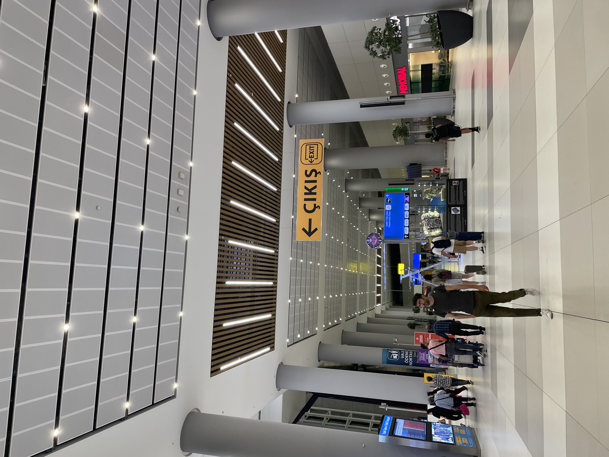 This is a photograph of a large, modern airport terminal interior in Arnavutköy, Turkey, likely Istanbul Airport given the signage. The space is characterized by a high, slatted wooden ceiling with embedded lighting, and large, grey cylindrical columns supporting the structure. The floor is tiled in a pattern of light and dark grey and white squares.
In the foreground, a man wearing a dark olive green t-shirt and dark olive green trousers walks away from the camera. He has glasses and a backpack. Further into the background, a group of people are walking in various directions. Some are pulling luggage, including a woman with a pink bag and a man with a trolley. Another man in a dark t-shirt and black shorts is seen walking away from the camera towards the right side of the frame.
Visible signage includes a yellow directional arrow sign with "ÇIKIŞ" (Exit in Turkish) and an English "EXIT" symbol. Other signs are present, including a blue digital display board on the left showing public transport information and a sign that reads "VALE ÖDEME NOKTASI" (Valet Payment Point). In the distance, there are shops with prominent signage such as "MEMORIAL". The overall ambiance is that of a busy international travel hub.