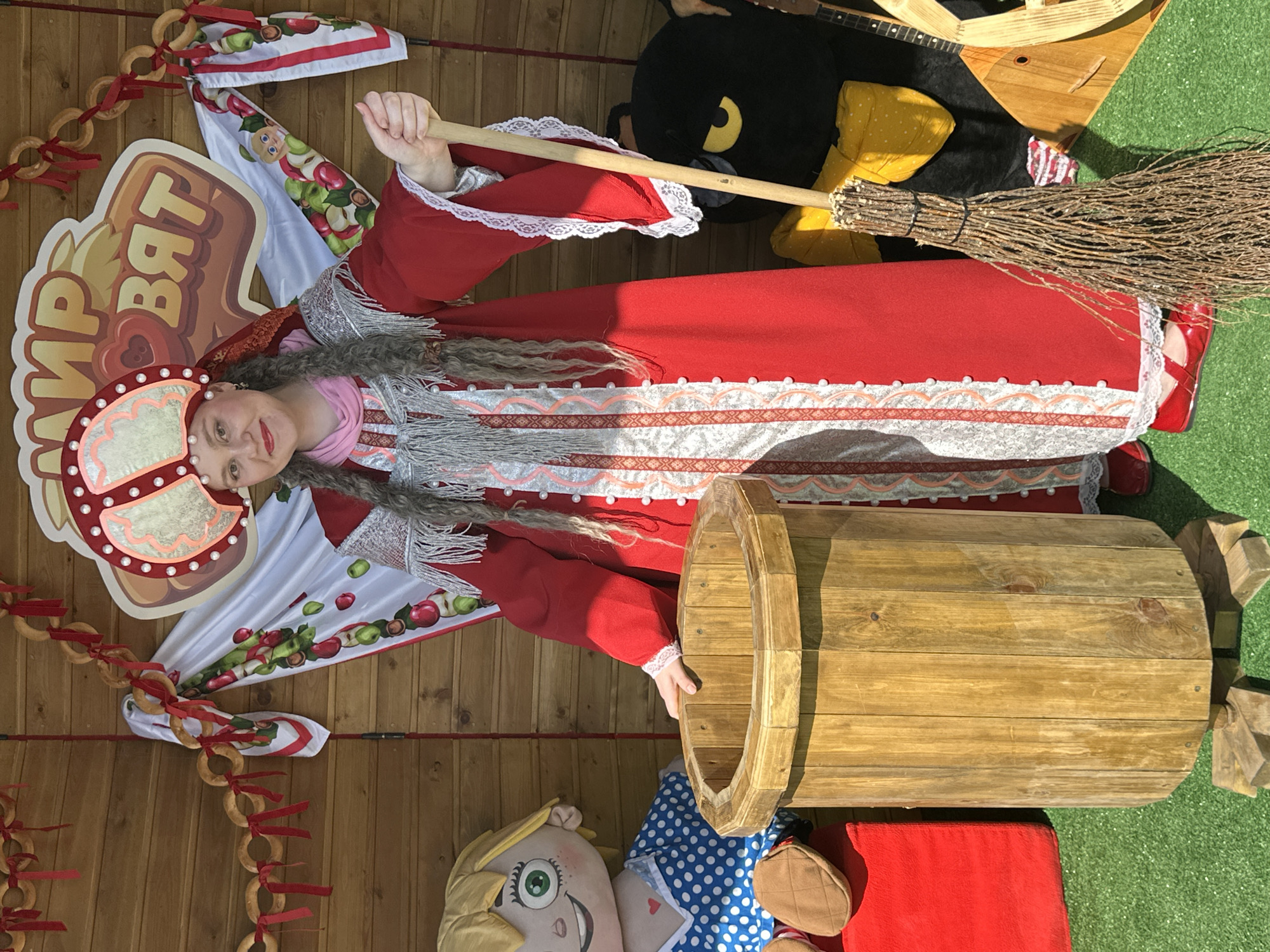 A woman in a traditional red dress and ornate headdress stands on a green, artificial turf ground in front of a wooden wall. She holds a broom made of twigs in her right hand. To her left is a large, wooden barrel, and to her right, partially obscured, is a black stuffed bird with yellow eyes and a balalaika. A large doll with blonde hair and a blue polka-dot dress is positioned to the left of the barrel. Behind the woman is a banner with the Cyrillic text "МИР ВЯТ". The overall scene appears to be set up for a festive or cultural event.