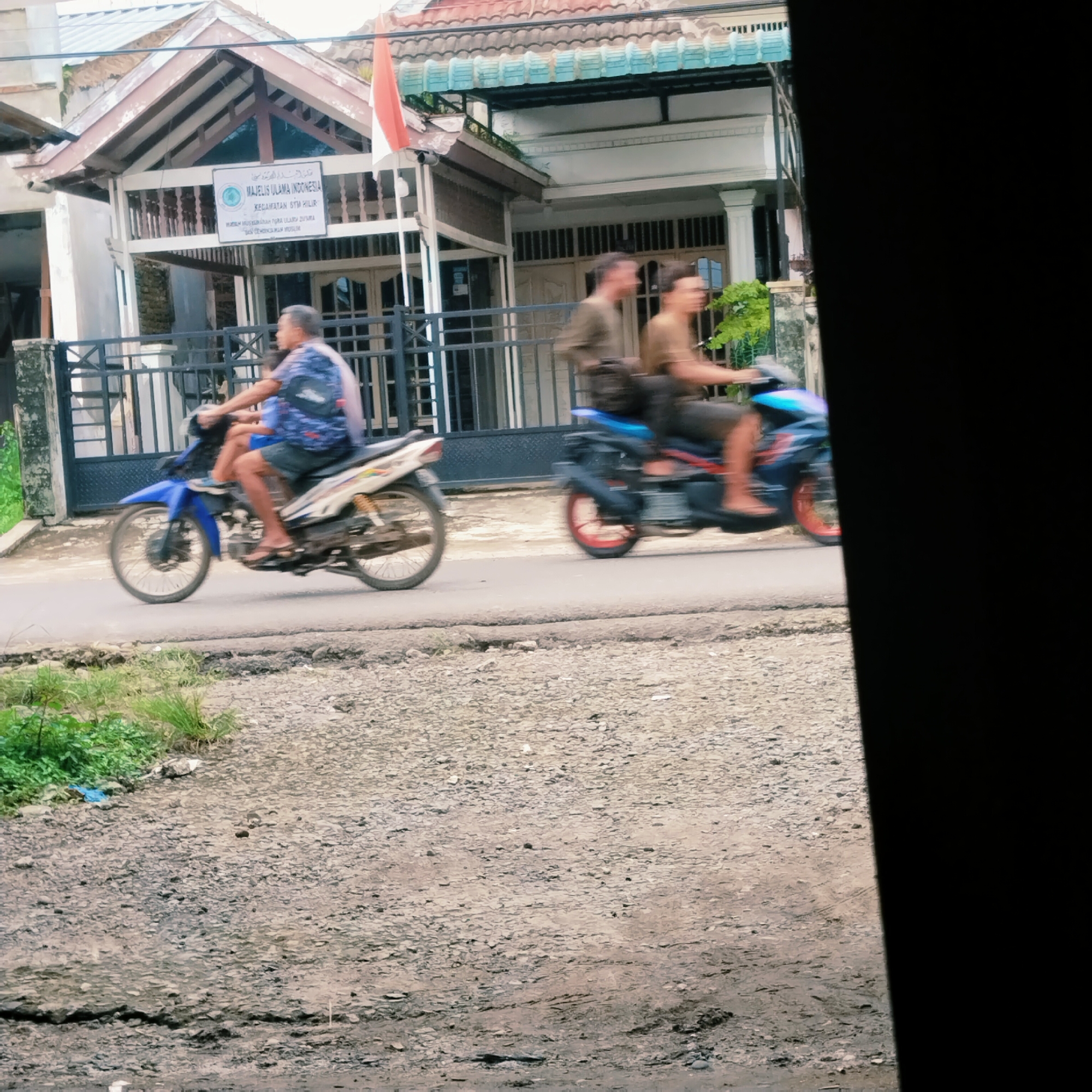 The image depicts a street scene in Talun Kenas, Indonesia, featuring two motorcycles in motion. In the foreground, a motorcycle travels from left to right, carrying two individuals. The rider, an older male with short dark hair, wears a blue patterned t-shirt and dark shorts. He is accompanied by a passenger, likely a child, seated in front of him.
Further down the street, a second motorcycle also moves from left to right. This motorcycle is ridden by two individuals. The rider, a male with short brown hair, wears a dark olive green t-shirt and dark shorts. The passenger, also a male with short brown hair, is seated behind the rider and appears to be wearing a dark olive green t-shirt and dark pants.
The background reveals a building with a prominent sign displaying "MAJELIS ULAMA INDONESIA" and "KECAMATAN" which suggests it is a local religious council or community building. An Indonesian flag, red and white, is visible on a flagpole near the building. The environment includes a paved road, a gravelly shoulder with some sparse vegetation, and a fence bordering the property. The overall lighting suggests an overcast day.