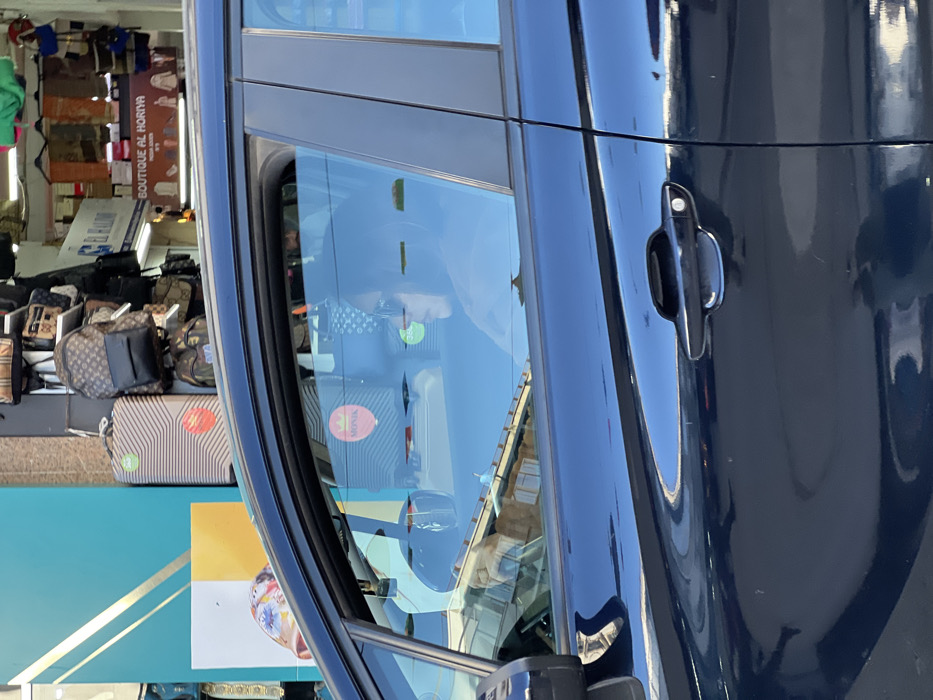 This image captures a static street scene in Tanger, Morocco, observed from the perspective of a vehicle. The foreground features the dark blue driver's side of a car, encompassing both the front and rear passenger doors, with reflections of the surroundings visible on its glossy surface and windows. Inside the vehicle, a person is seated in the driver's seat, seen in profile looking towards the right. They wear dark sunglasses and have dark hair, with hands positioned near the steering wheel.
Beyond the car, the background reveals a commercial street. To the left, a vibrant teal-colored wall or shop facade with gold diagonal stripes is partially visible. Immediately to its right, a retail shopfront displays an array of merchandise, including several backpacks with distinctive patterns resembling luxury brands (e.g., Louis Vuitton monogram and Gucci designs), various handbags, and a beige striped hard-shell suitcase featuring a green circular sticker. Prominent signage above the shop includes "BOUTIQUE AL HORIYA" (Liberty Boutique) in both French and Arabic, with another partially visible Arabic sign above it that reads "EL HALOUF". A "MONIK" logo is also visible as a reflection within the car's window. The scene is set on an urban thoroughfare in Tanger.