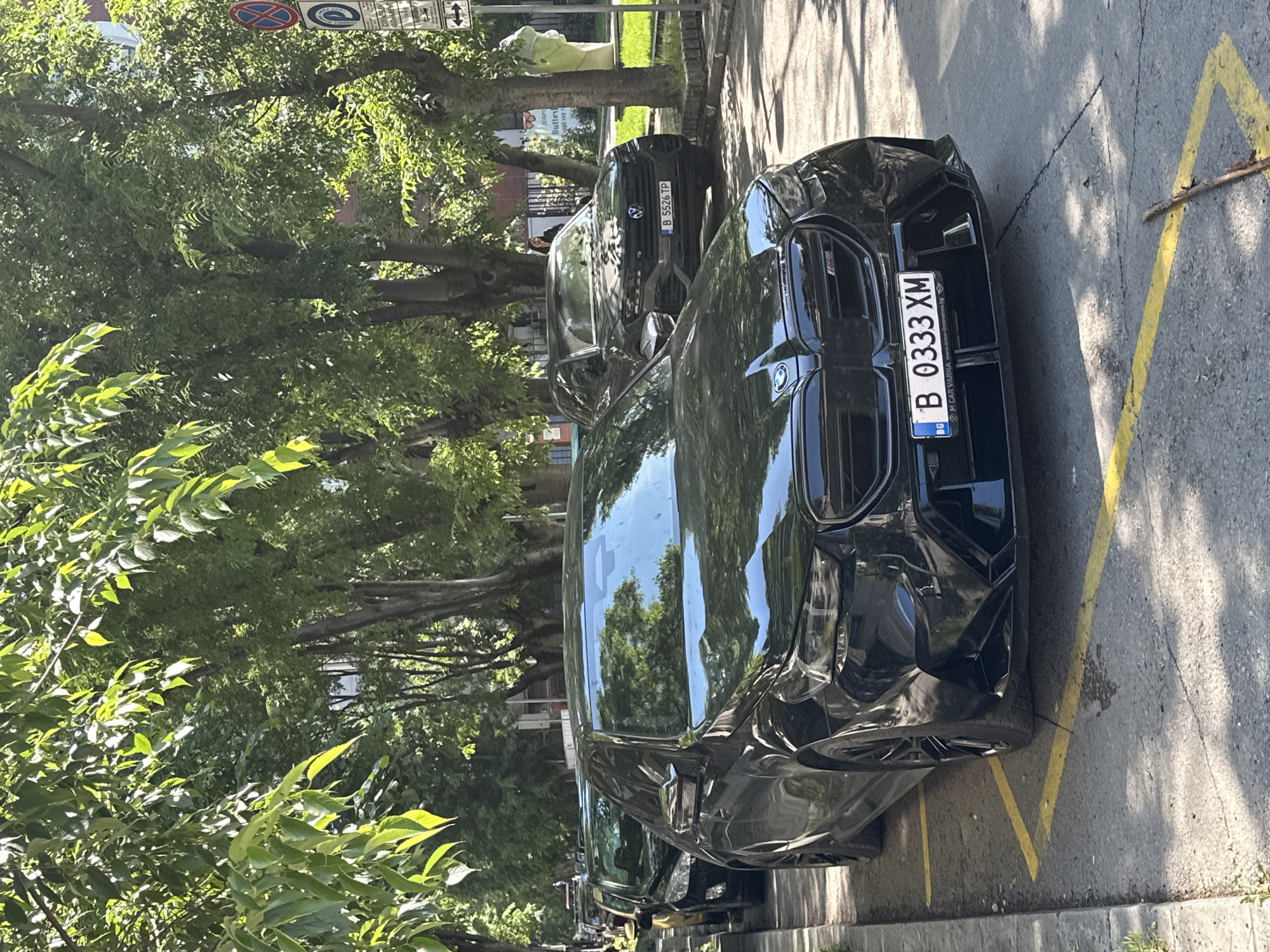 The image shows two cars parked on a city street. In the foreground, there is a black BMW M series car, with a license plate reading "B 0333 XM" from Varna, Bulgaria. The license plate also has the website "mcaryana.bg" printed. Behind the BMW is a dark-colored SUV, with a license plate reading "B 5526 TP". The cars are parked next to a curb with yellow parking lines on the pavement. The area is lined with trees, and a street sign is visible in the upper left corner of the frame. There is a business sign in Cyrillic visible behind the SUV. The environment is sunny and appears to be a residential or commercial area in Varna, Bulgaria.