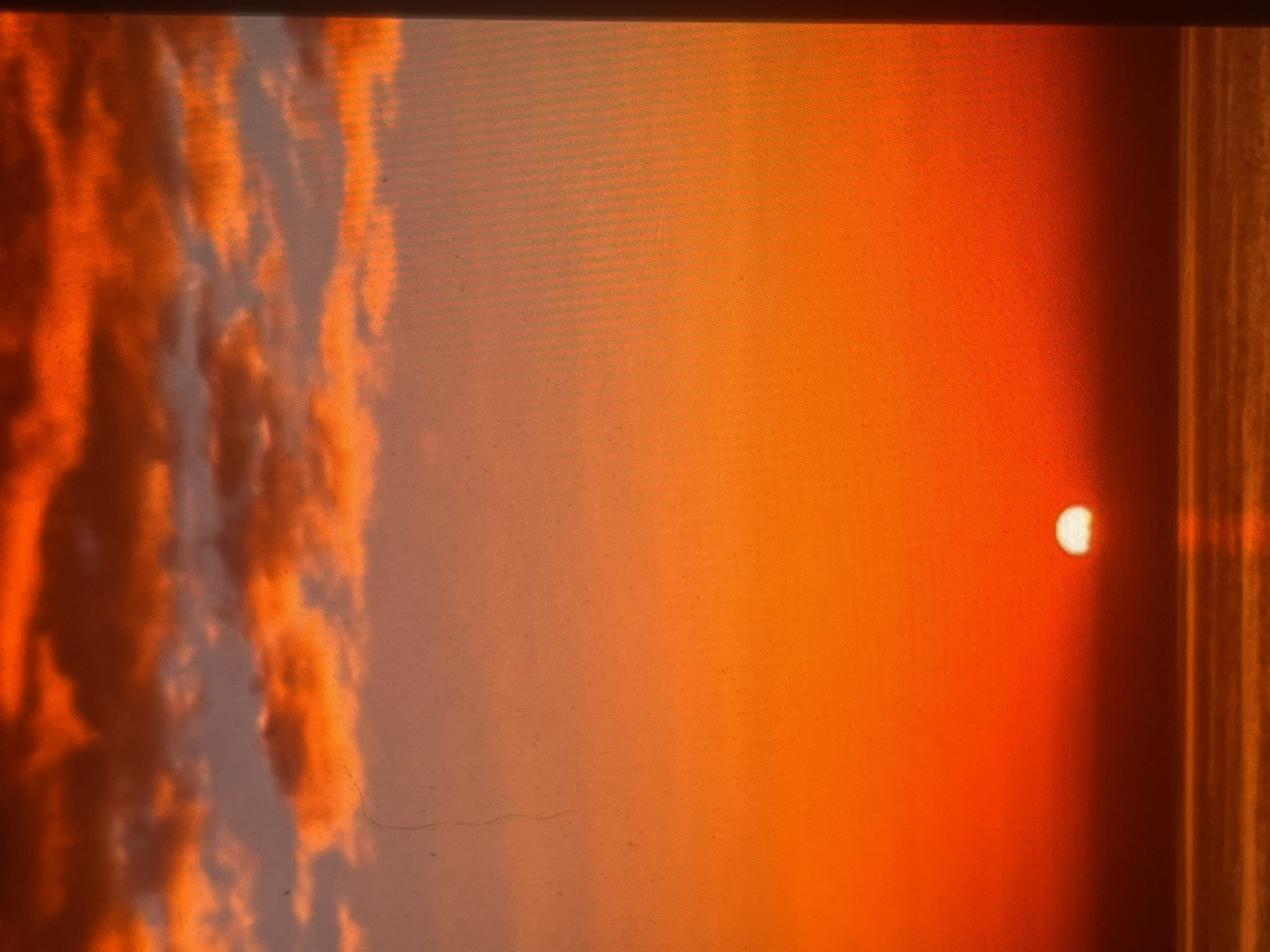 The image captures a vibrant sunset over an expanse of water, with the scene located in Zelenyi Hai, Ukraine. The sky presents a striking gradient of reds and oranges, deepening to fiery red at the zenith and transitioning to brighter orange near the horizon. Multiple layers of dark orange and red clouds are positioned in the upper sky, interspersed with areas of lighter grey-blue. A brilliant, white-yellow semi-circular sun is visible at the horizon line in the lower right, partially submerged below the water's surface and casting intense light. The body of water below reflects the sky's colors, appearing in rich shades of orange and red, with subtle texture suggesting calm ripples. No human figures, vessels, or specific geographical landmarks are discernible. Forensic examination reveals a pronounced diagonal grid pattern, indicative of a digital screen or a low-resolution scan, along with various dust particles and a faint vertical scratch across the image surface.