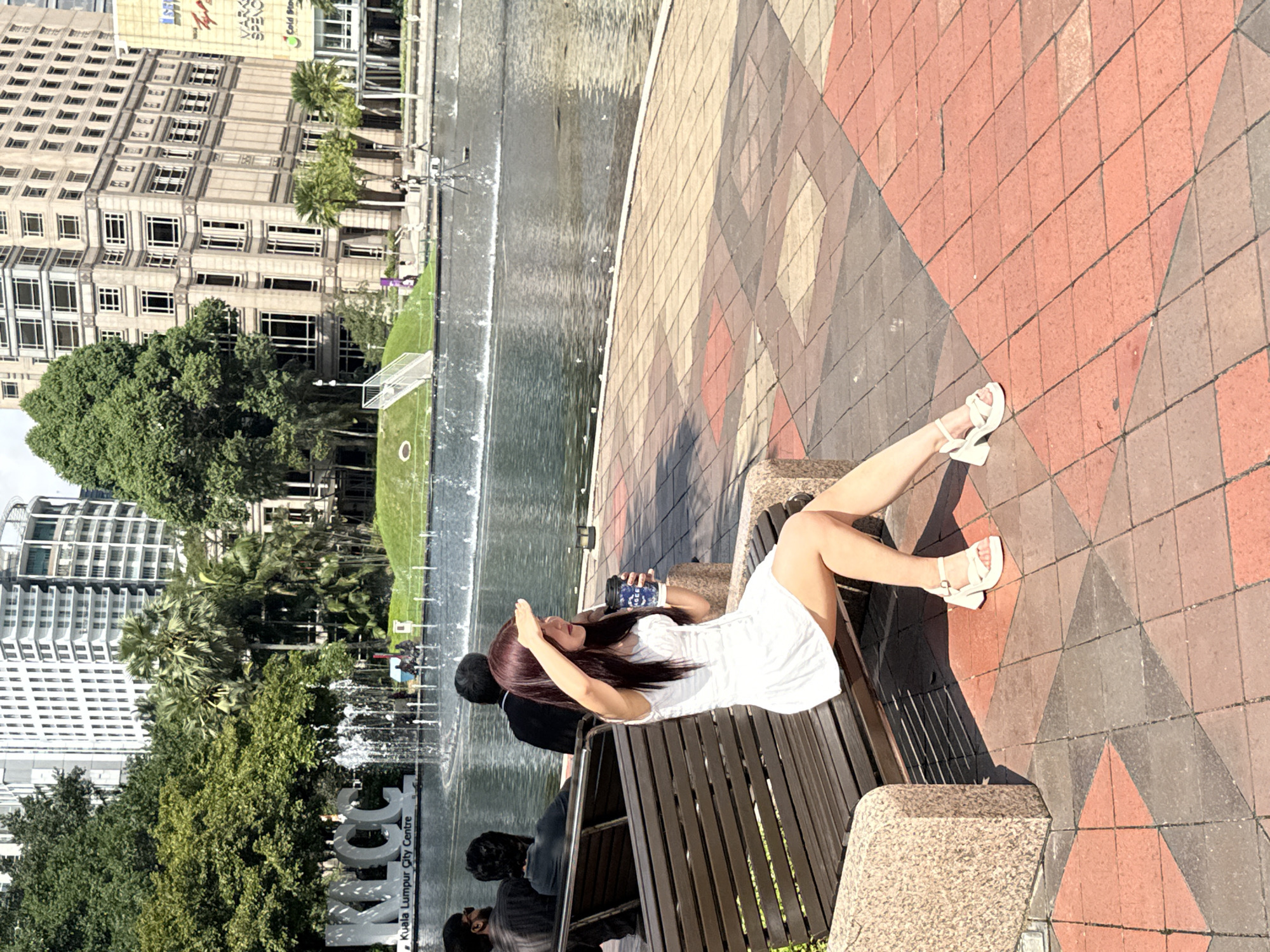 Here is a detailed description of the image:
The image is shot from a ground-level, medium distance, and depicts a woman sitting on a park bench in Kuala Lumpur City Centre (KLCC), Malaysia.
The woman, who appears to be young, is sitting on the bench with her legs extended towards the right side of the frame. She is wearing a white dress and white sandals. Her dark hair cascades down her shoulders. In her hand, she holds a dark blue container with a white logo. With her other hand, she is shielding her eyes from the sun.
Other individuals are sitting on the same bench, though facing away from the camera. A man with black hair in a black suit jacket is sitting just to her left, and another individual with their back to the camera can be seen.
Behind the bench is a rectangular fountain. Beyond the fountain are trees, buildings, and a well-maintained green lawn with a soccer goal, revealing the setting as the KLCC park in Kuala Lumpur. The buildings in the backdrop contribute to the urban scenery.
The ground surrounding the bench is paved with patterned tiles in shades of red, gray, and beige. The image seems to be taken during daylight, with strong sunlight casting shadows on the scene.