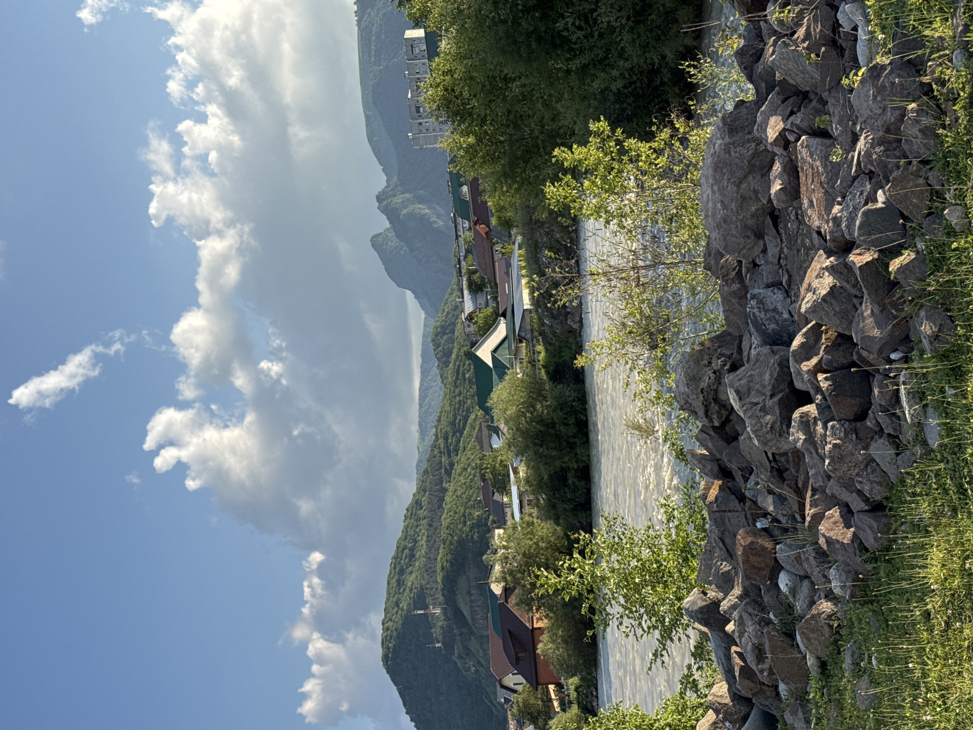 The image depicts a wide, fast-flowing river in the foreground, bordered by a rocky embankment. Beyond the river, a small town or village is nestled amongst lush green hills and mountains. Numerous houses with red and brown tiled roofs are visible, interspersed with trees. In the distance, larger mountains rise, with some appearing rocky and others covered in vegetation. The sky above is a bright blue with scattered white clouds, casting dappled sunlight on the landscape. A tall, slender structure, possibly a radio tower or pylon, is visible on one of the hills. The overall scene is one of natural beauty with human settlement integrated into the mountainous terrain. The location is the city of Karachayevsk, Russia.