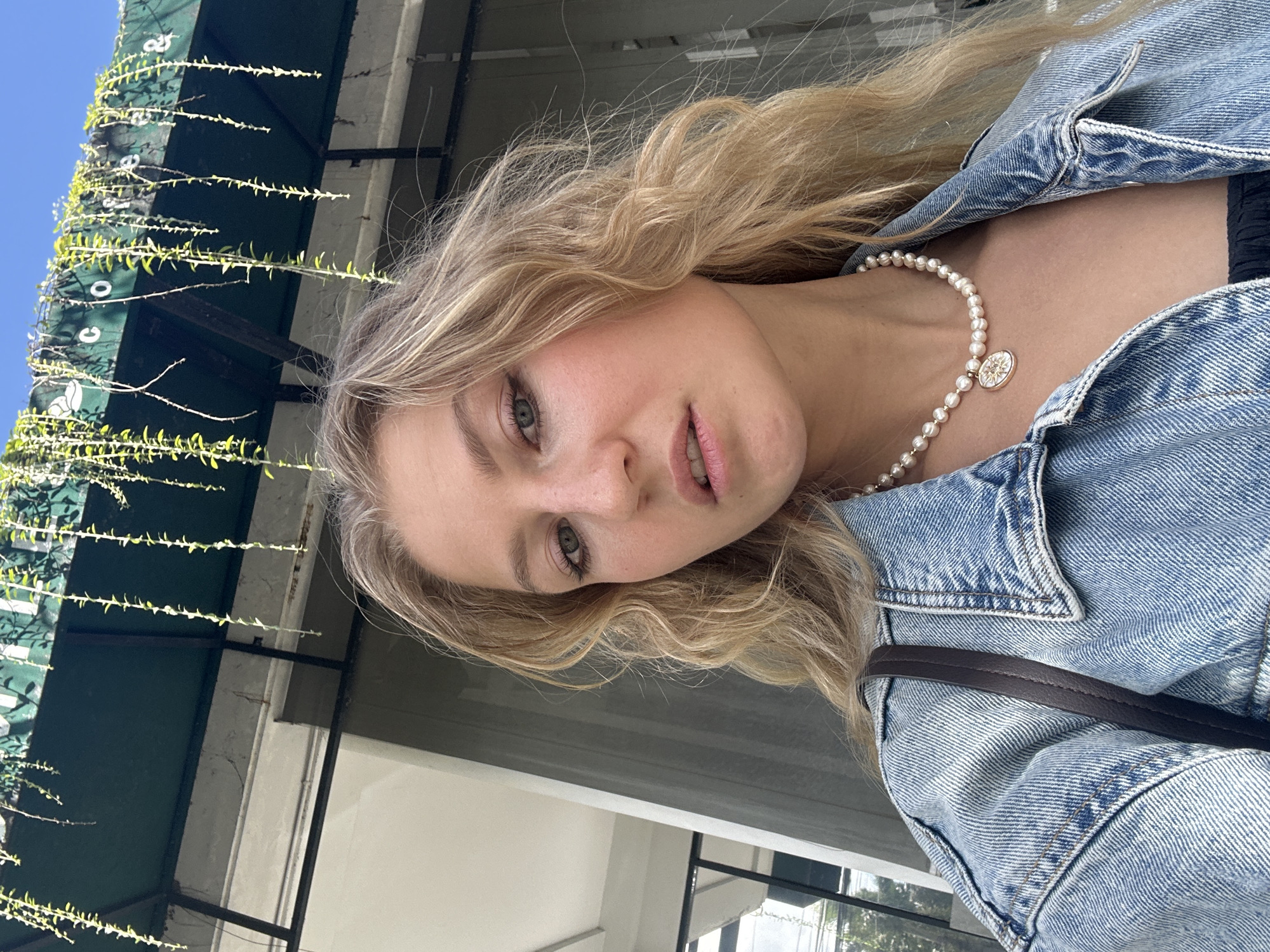 A young woman with blond wavy hair is posing for a selfie outdoors. She is wearing a denim jacket and a pearl necklace with a gold pendant. A dark-colored strap of a bag is visible over her right shoulder. Behind her, there's a building facade with a green awning covered in climbing plants. The sky is visible in the upper left corner, indicating it is daytime. The location appears to be an urban area, but the specific details to pinpoint Kuta Selatan, Indonesia, are not evident in the image.
