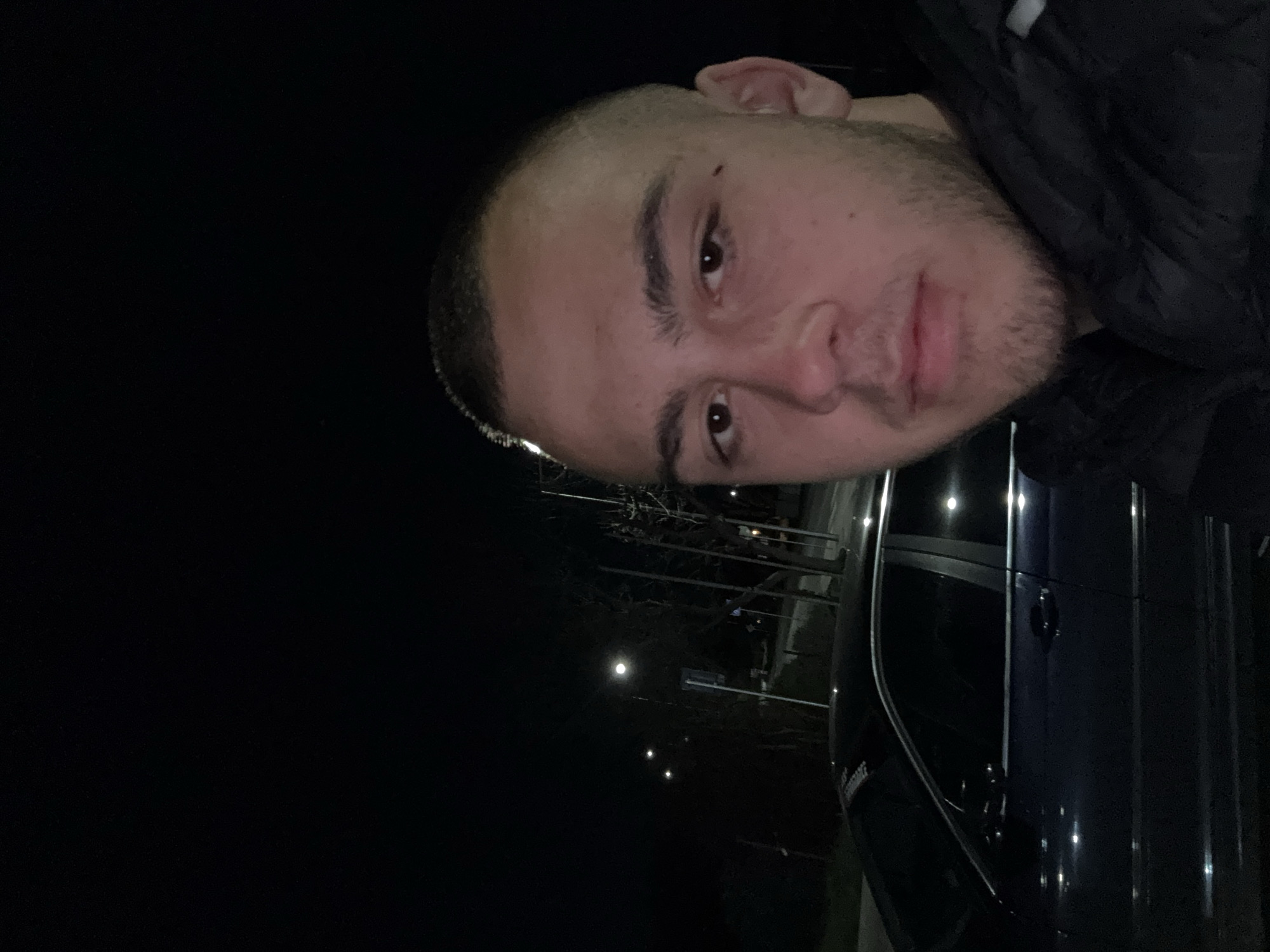 A man with closely cropped hair and a beard is visible in the foreground. He is wearing a dark jacket. To his right is a dark vehicle with a visible door handle. In the background, several street lights are visible against a dark sky. The location is potentially Topoli, Bulgaria.