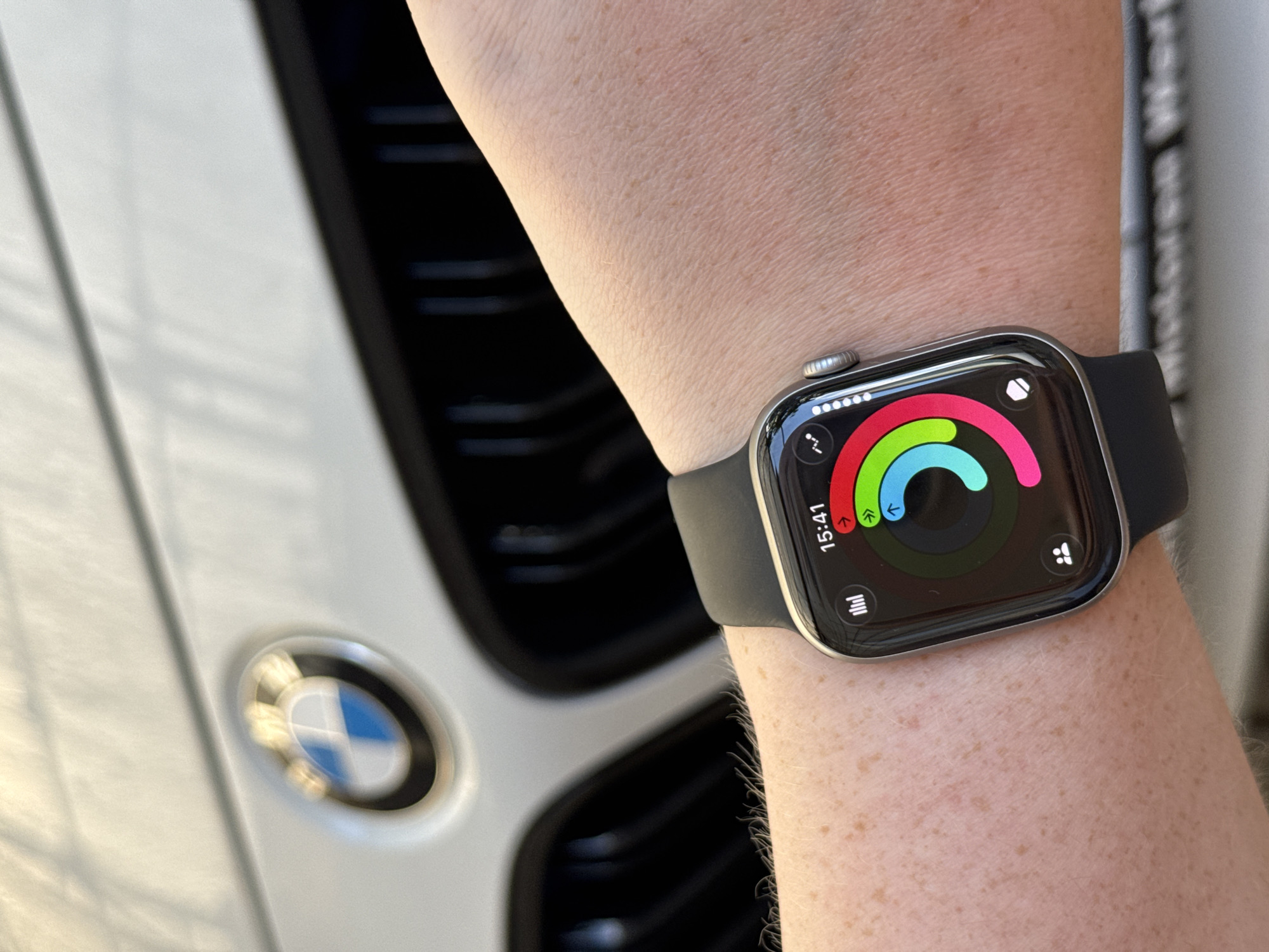 The image shows a close-up of a person's wrist wearing a smartwatch in front of a BMW car grille. The smartwatch displays activity rings, indicating goals for closing rings, exercising, and standing. The time on the watch reads 15:41. The background is blurred, but the distinctive BMW logo is visible on the car. The image does not provide information about the specific location in Mikhaylovsk, Russia.