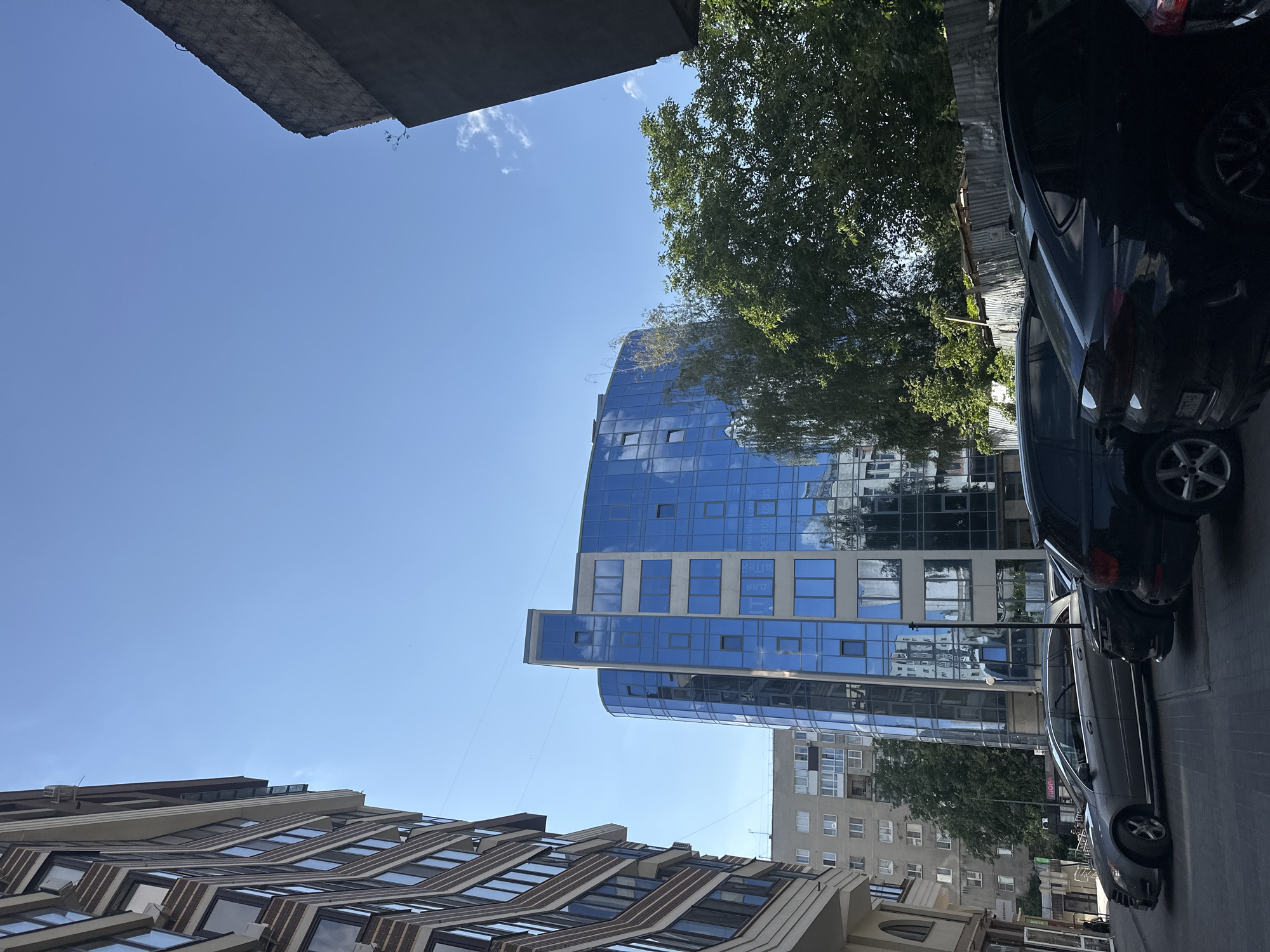 The image depicts a street scene in Ternopil, Ukraine, characterized by modern architecture and parked vehicles under a clear blue sky. On the left, a multi-story building with a textured facade and numerous windows occupies the foreground. To its right, a strikingly modern, curved building with a facade dominated by reflective blue glass stands prominently. The blue glass reflects the surrounding buildings and the sky. Further in the background, a light-colored residential building with multiple windows is visible, partially obscured by trees.
In the foreground, a row of parked cars is lined up along the street. The cars are of various makes and models, primarily dark in color, with one lighter-colored sedan positioned towards the left. The street itself appears paved with cobblestones or similar material. Dense green trees are interspersed between the buildings and behind a corrugated metal fence, adding a natural element to the urban landscape. The overall impression is of a well-maintained urban environment on a sunny day.