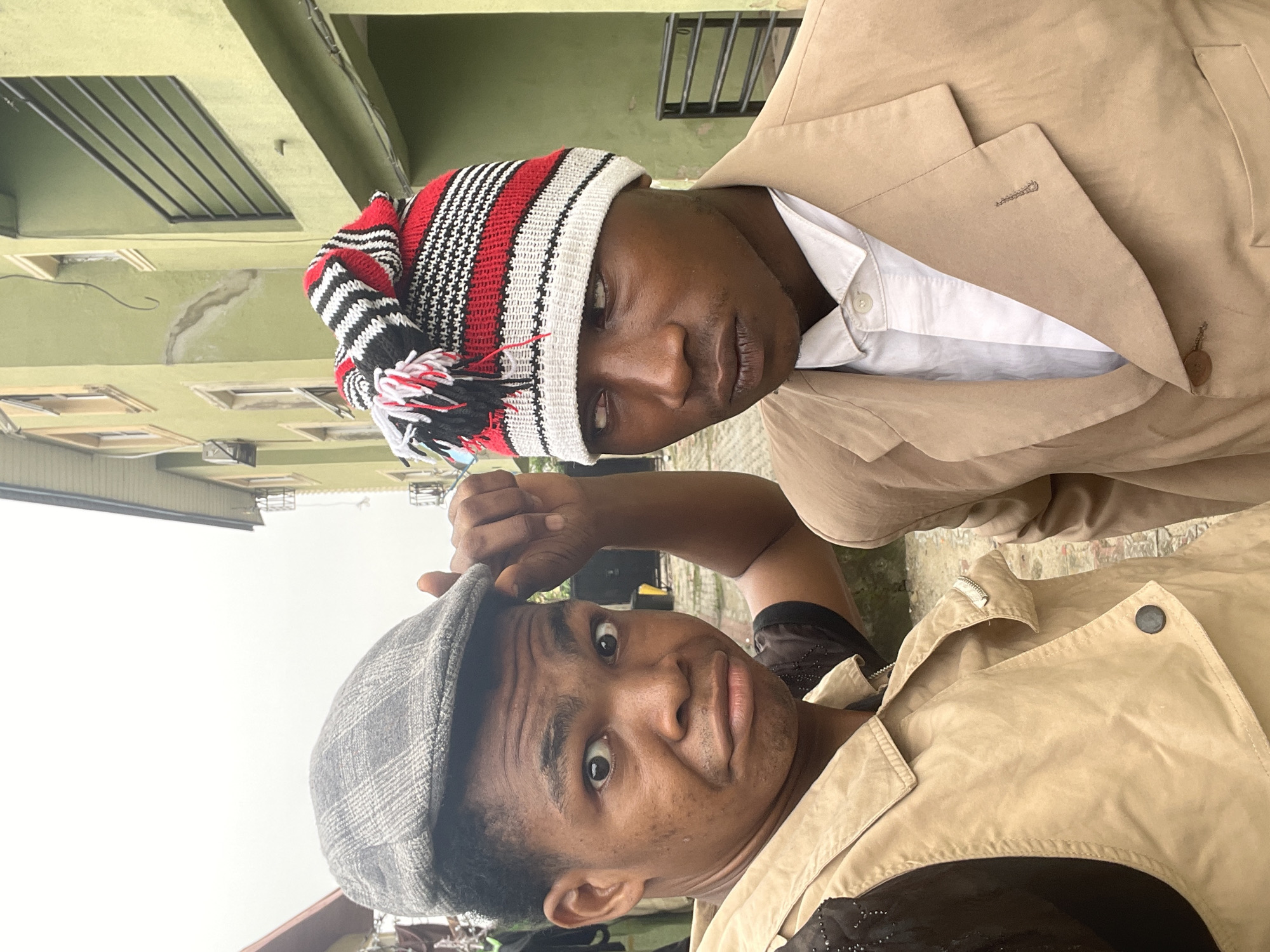 Two men are posing for a selfie outdoors. One man wears a red, white, and black knit hat and a beige blazer over a white shirt. The other man wears a gray flat cap and a tan jacket. They are in front of a light green multi-story building. The location is likely Ado, Nigeria, based on the prompt. The sky is overcast.